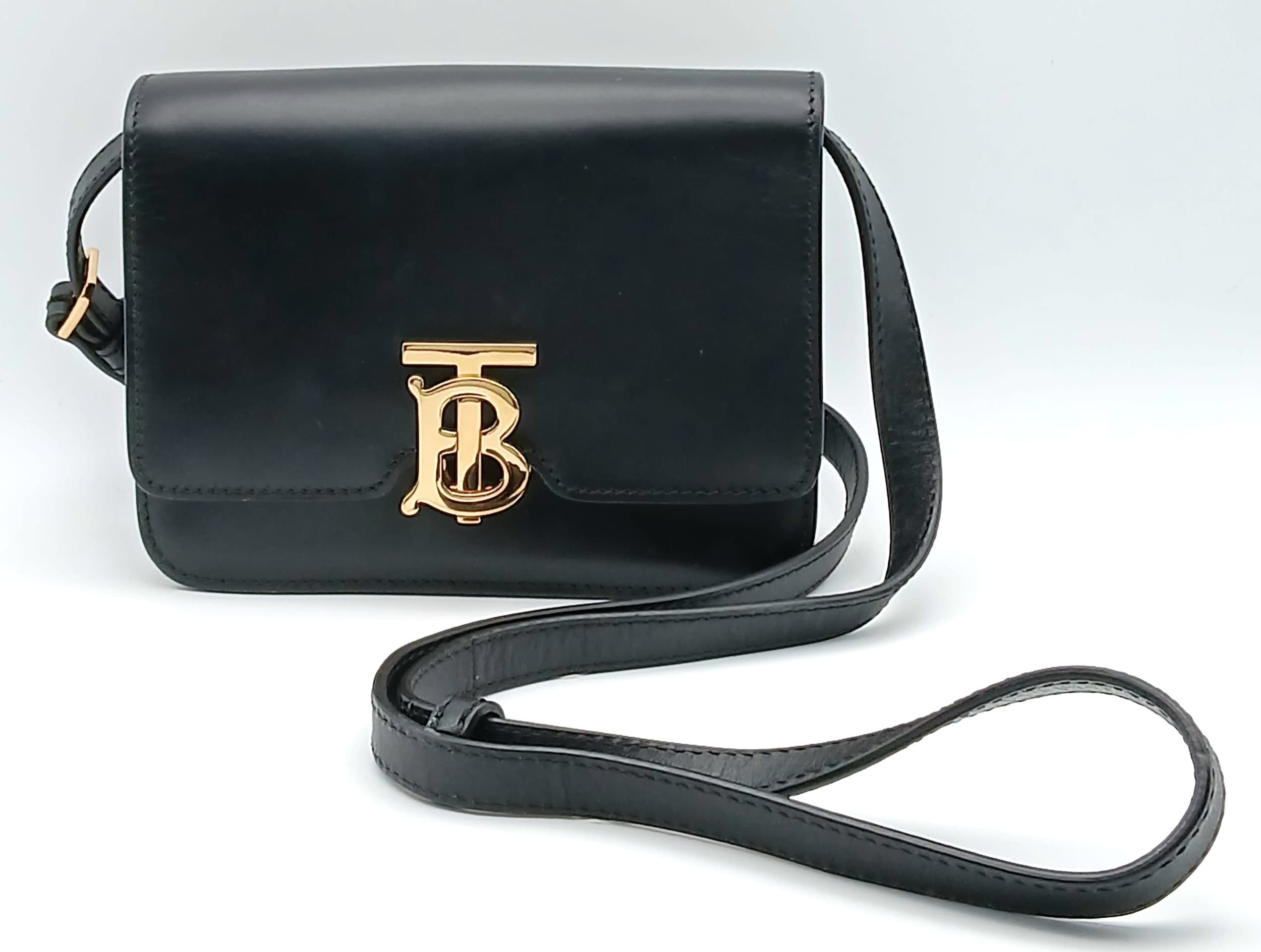 A Burberry Black Leather TB Crossbody Bag. Smooth black leather exterior with gold-tone hardware, adjustable shoulder strap, and front flap with signature TB monogram clasp. Black leather interior with two main compartments and an open inner side pocket. Comes with a dust cover bag and booklet. Approximately 17cm length x 12.5cm height x 5cm depth. In good overall condition with light surface scratches – please see photos or request a condition report. Ref: 27737