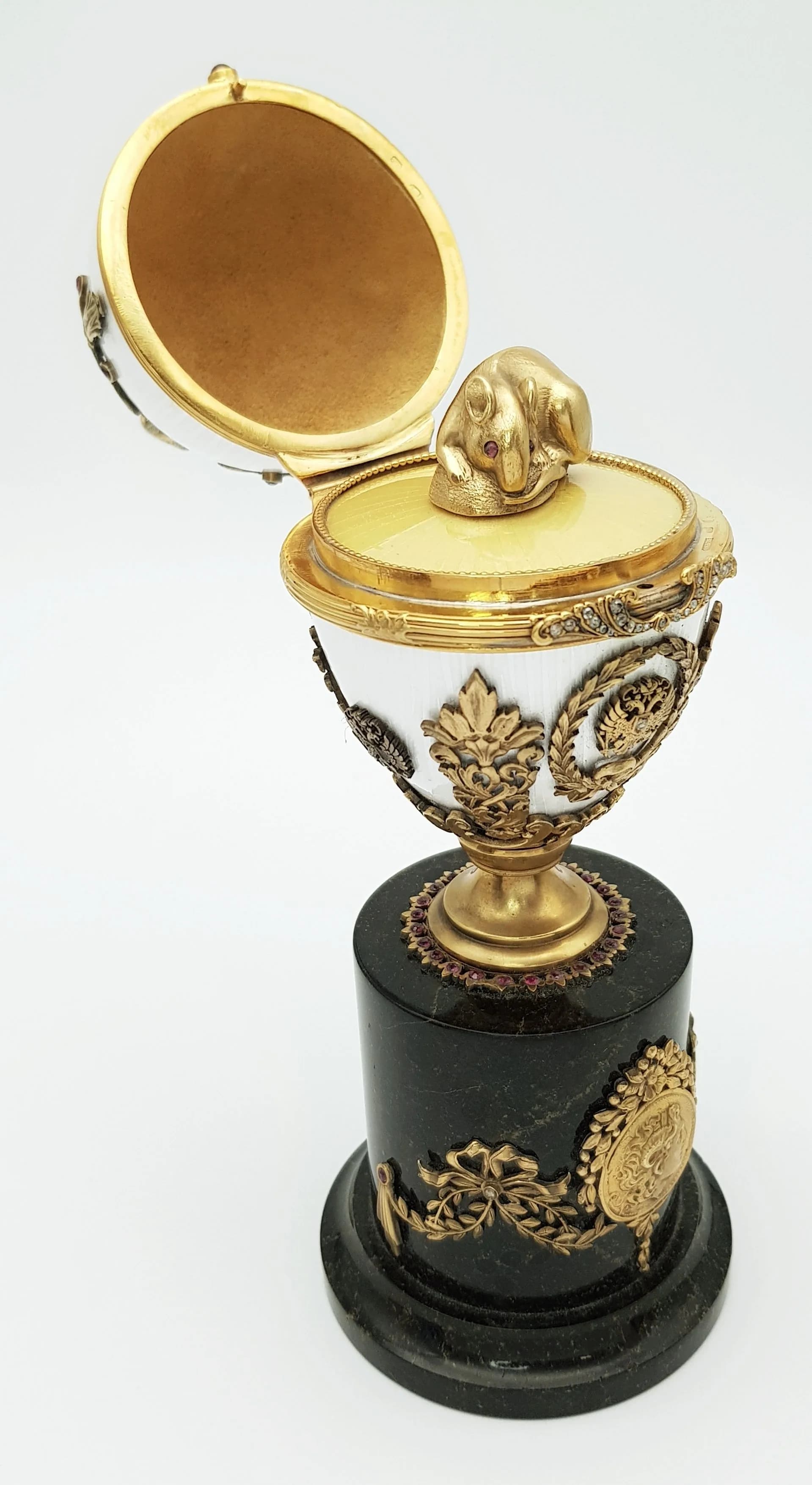 A Russian Style Diamond Set Gilded Silver and Enamel Surprise Egg. Russian Imperial style ‘Easter Egg’ with ovoid silver-gilt mounted body in white guilloche enamel, decorated with a diamond set 1913 motif, imperial eagle mounts, ribbon and laurel wreath detailing, and red stone accents. Opens to reveal a naturalistic silver mouse figure with purple gem set eyes. Raised on a black hardstone column base with further gilded decoration. Approx 19.9cm height. Total weight 580.7g. Comes with a presentation box. UK Mainland Shipping Only or arrange a courier service. Collection from Cadmore HQ is strongly advised.
