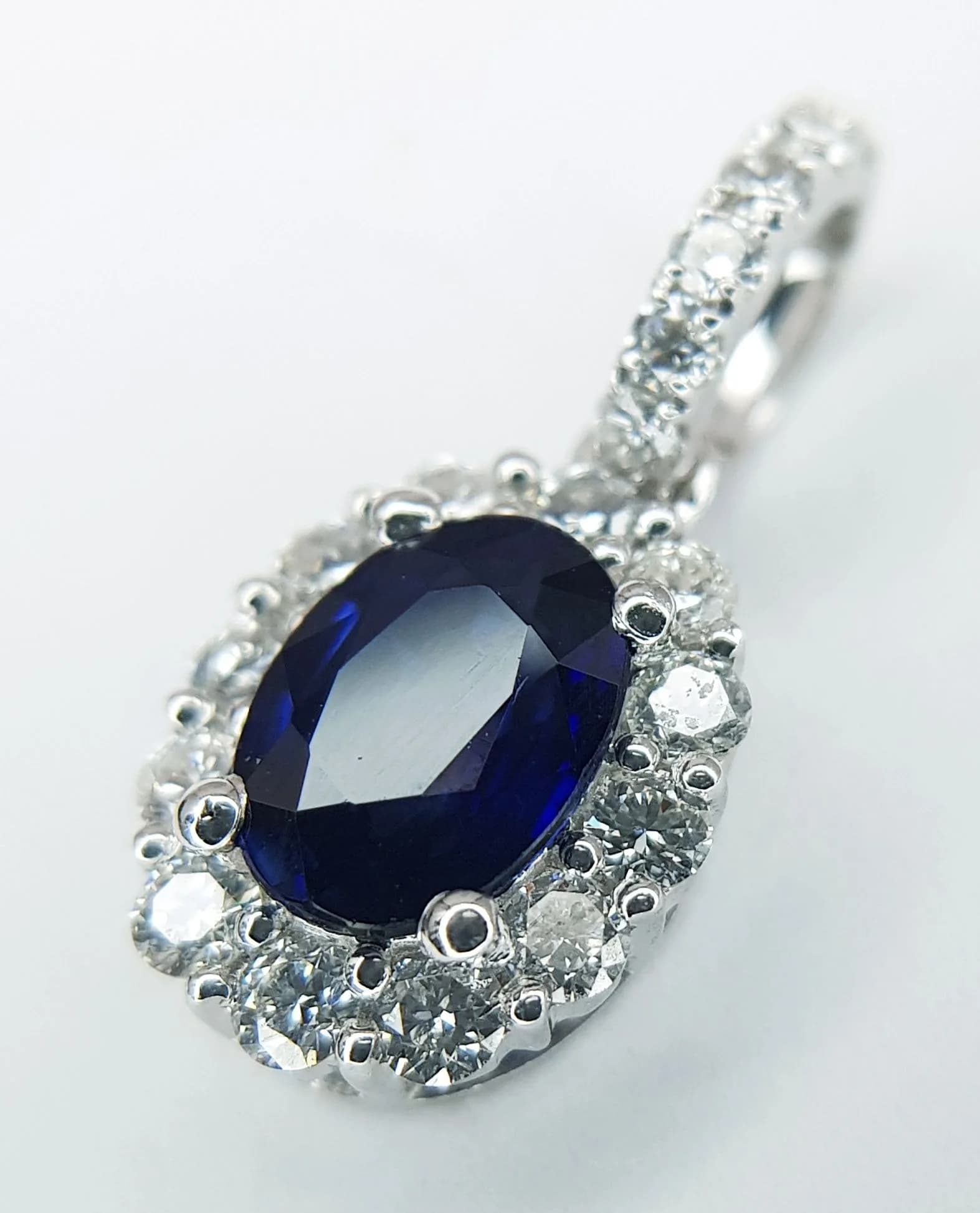 An 18K White Gold, Sapphire and Diamond Pendant. c.0.75ct oval cut sapphire with a round brilliant cut diamond surround. Diamond accents on bail. 15mm. 1.55g total weight. MT43.