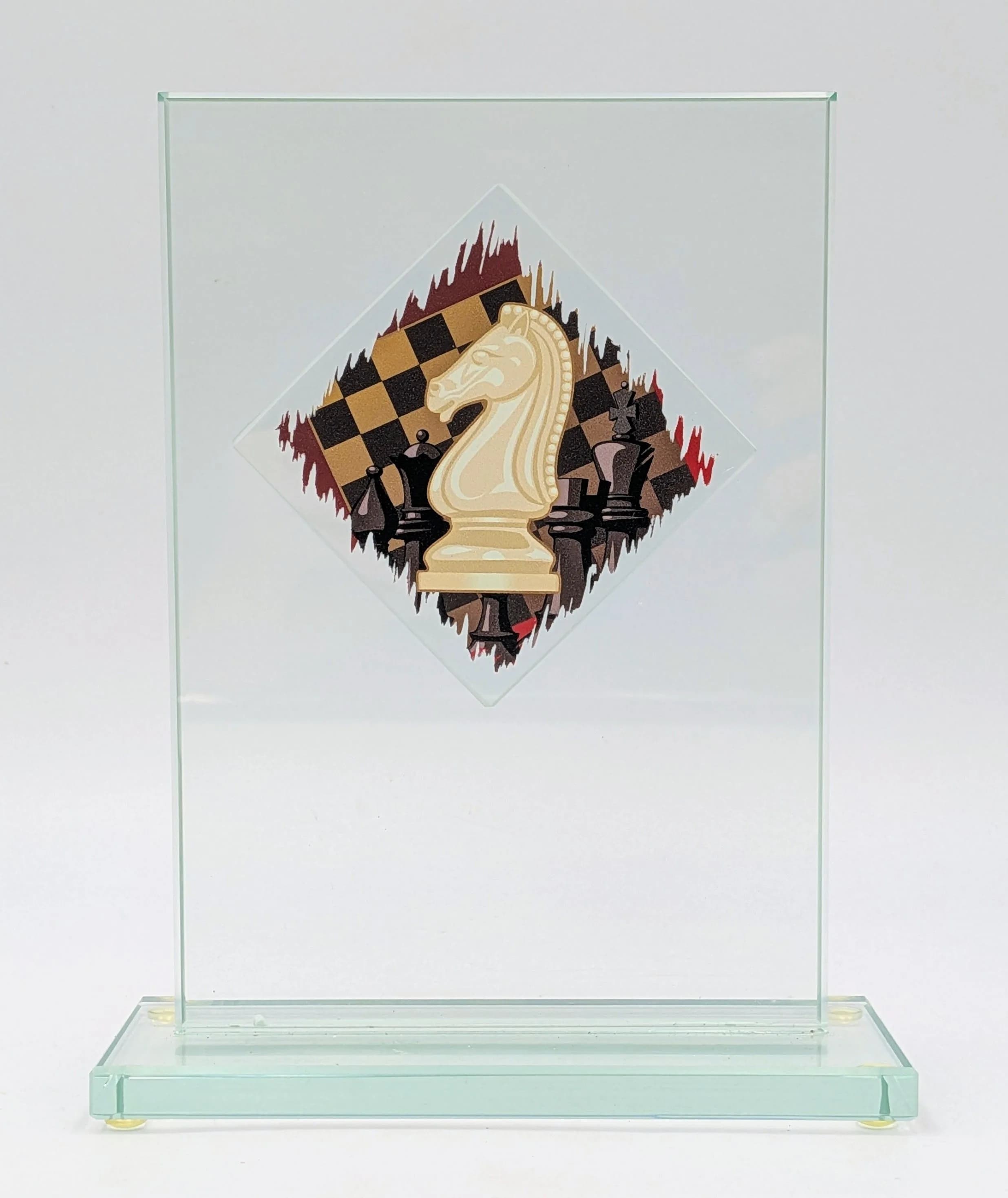 A Glass Chess Trophy. Featuring a central chess knight design with chess board and pieces decoration, on a clear glass base. Approx 19cm height including base x 12.5cm width. Base approx 15cm x 6cm.