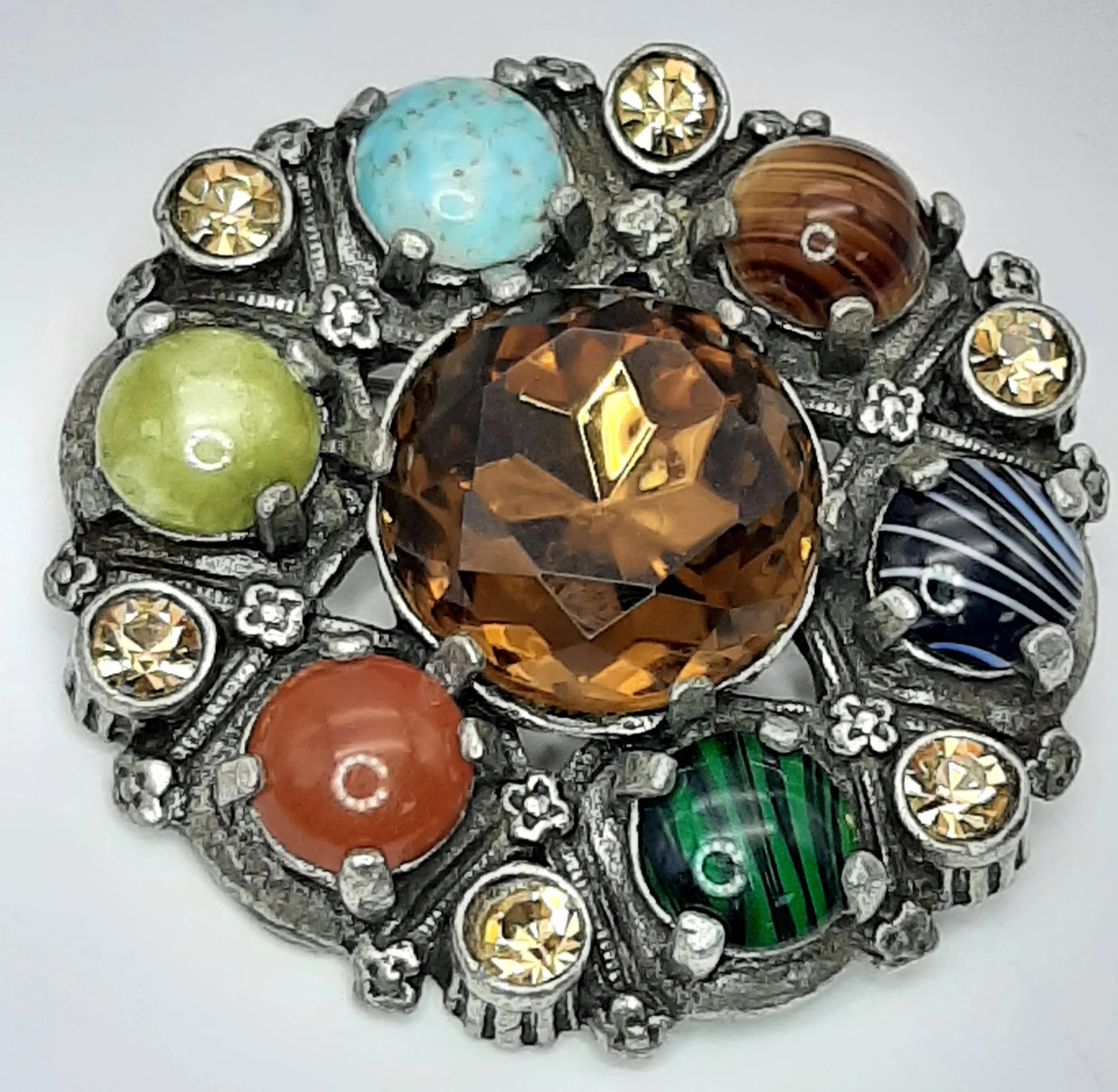 A Pretty Multi-Gemstone Brooch. Features a silver-tone circular design set with a large central smoky quartz stone, surrounded by turquoise, carnelian, malachite, tiger’s eye, agate, serpentine and citrine faceted stones. The brooch measures 5cm diameter.