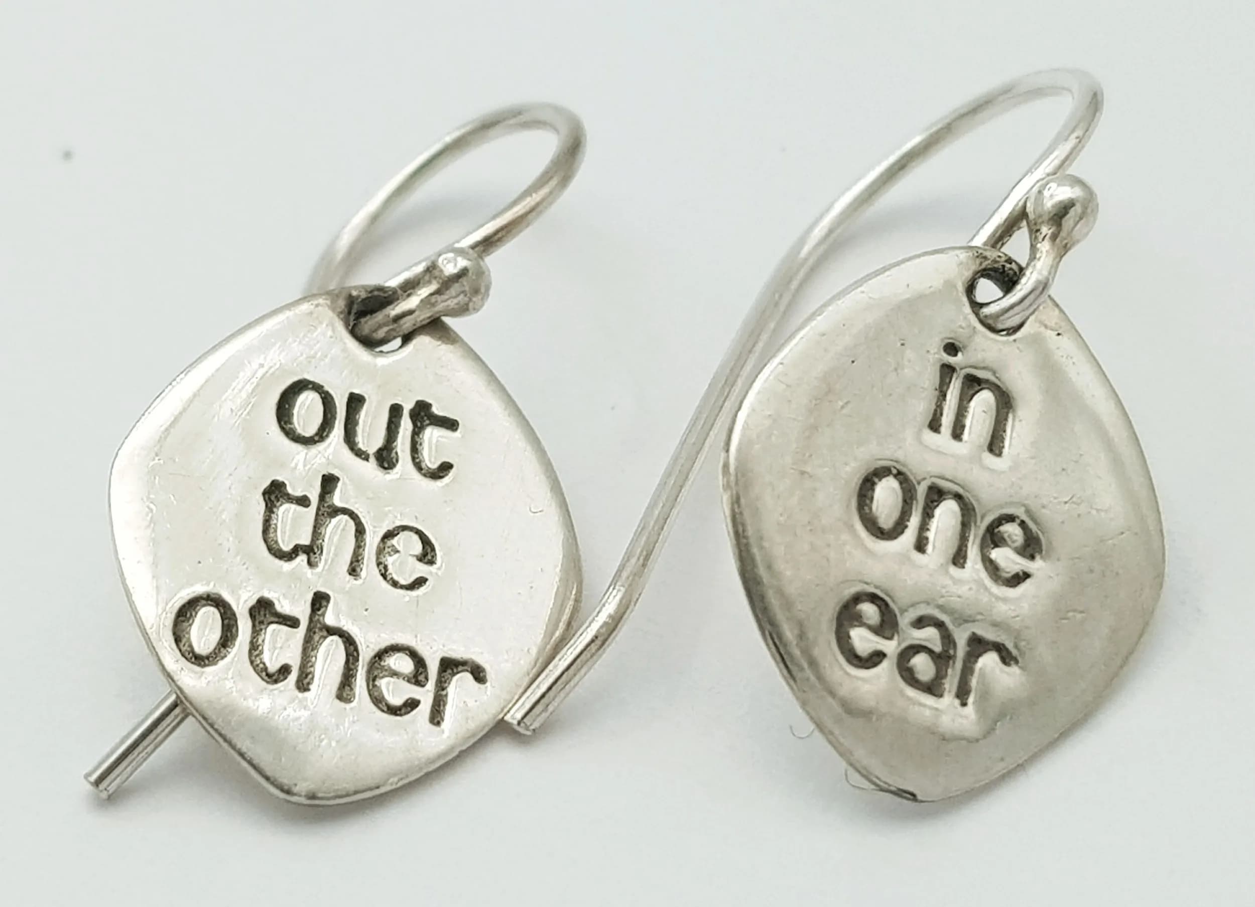 A Quirky Pair of Silver Guitar Pick Drop Earrings. Features guitar pick-shaped drops inscribed “out the other” and “in one ear”, with fish hook fittings. Each drop measures 1.1cm x 1.3cm. Gross weight 2.15 grams.