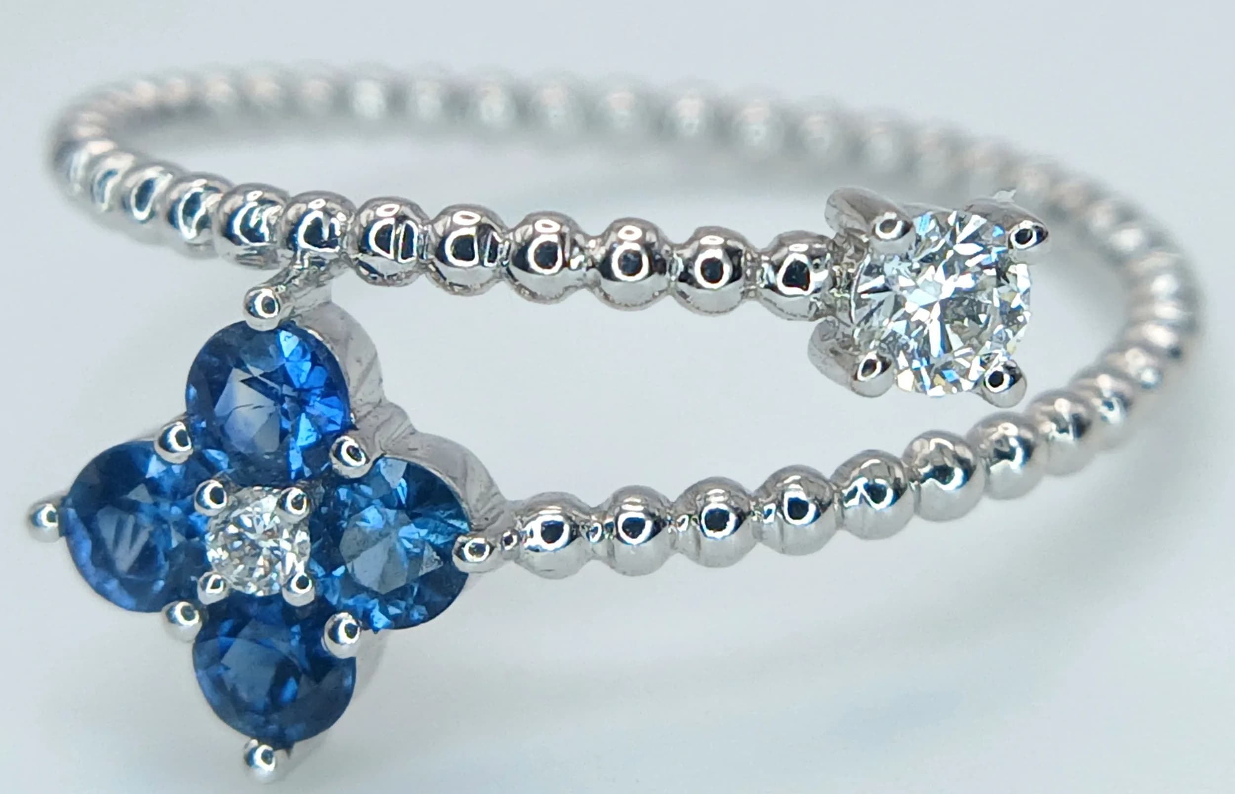 AN 18K WHITE GOLD, DIAMOND AND SAPPHIRE SET, FLOWER DESIGN BEADED CROSSOVER RING. 0.33CTW SAPPHIRES AND 0.12CTW DIAMONDS. 1.5G, SIZE M. SSPA 5001