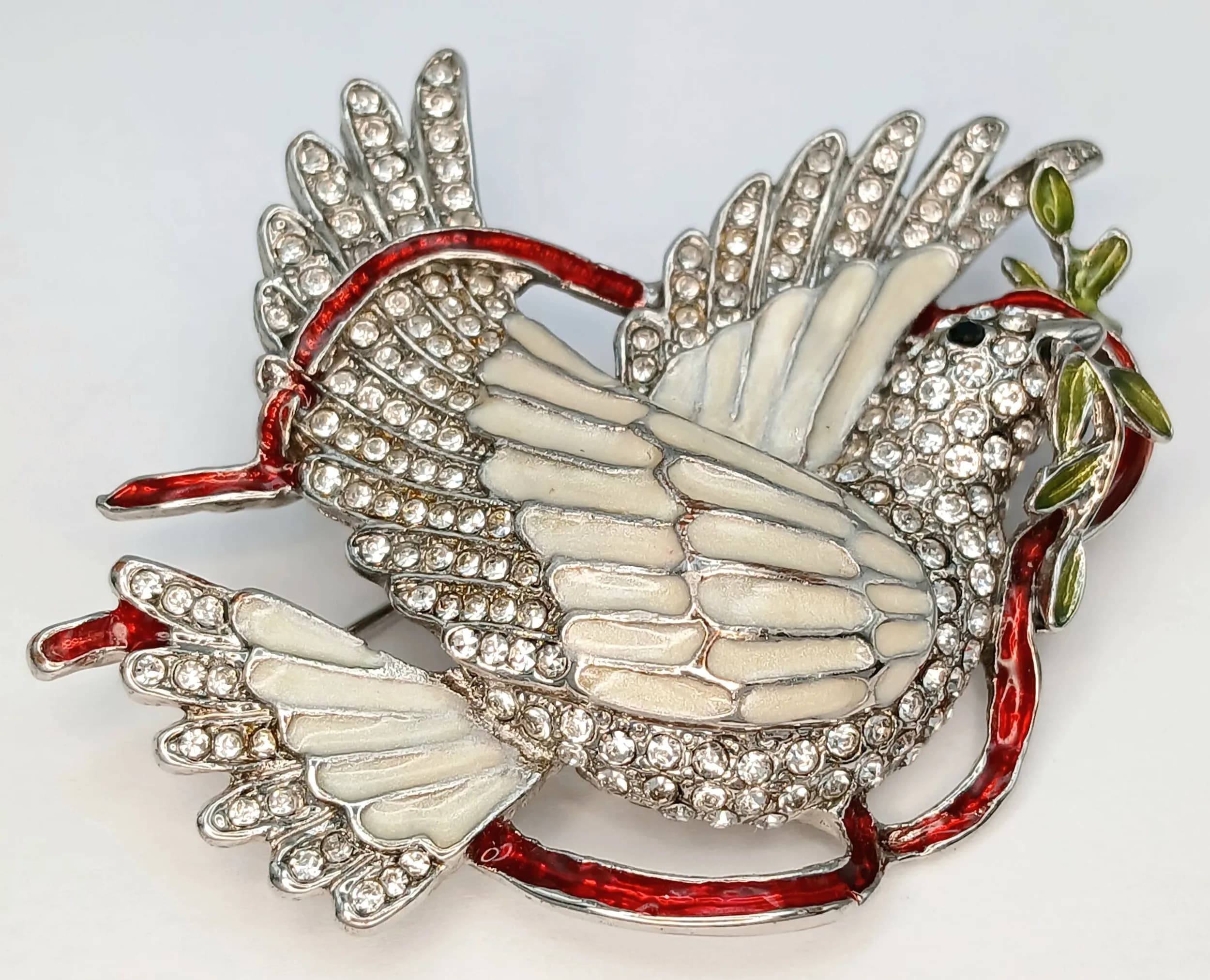 A Stunning Silver Tone Enamel and Crystal Set Dove Brooch. Features a white enamelled dove with outstretched wings, pavé set clear crystals, red enamel ribbon and green enamel olive branch details. The brooch measures 6cm x 4cm.