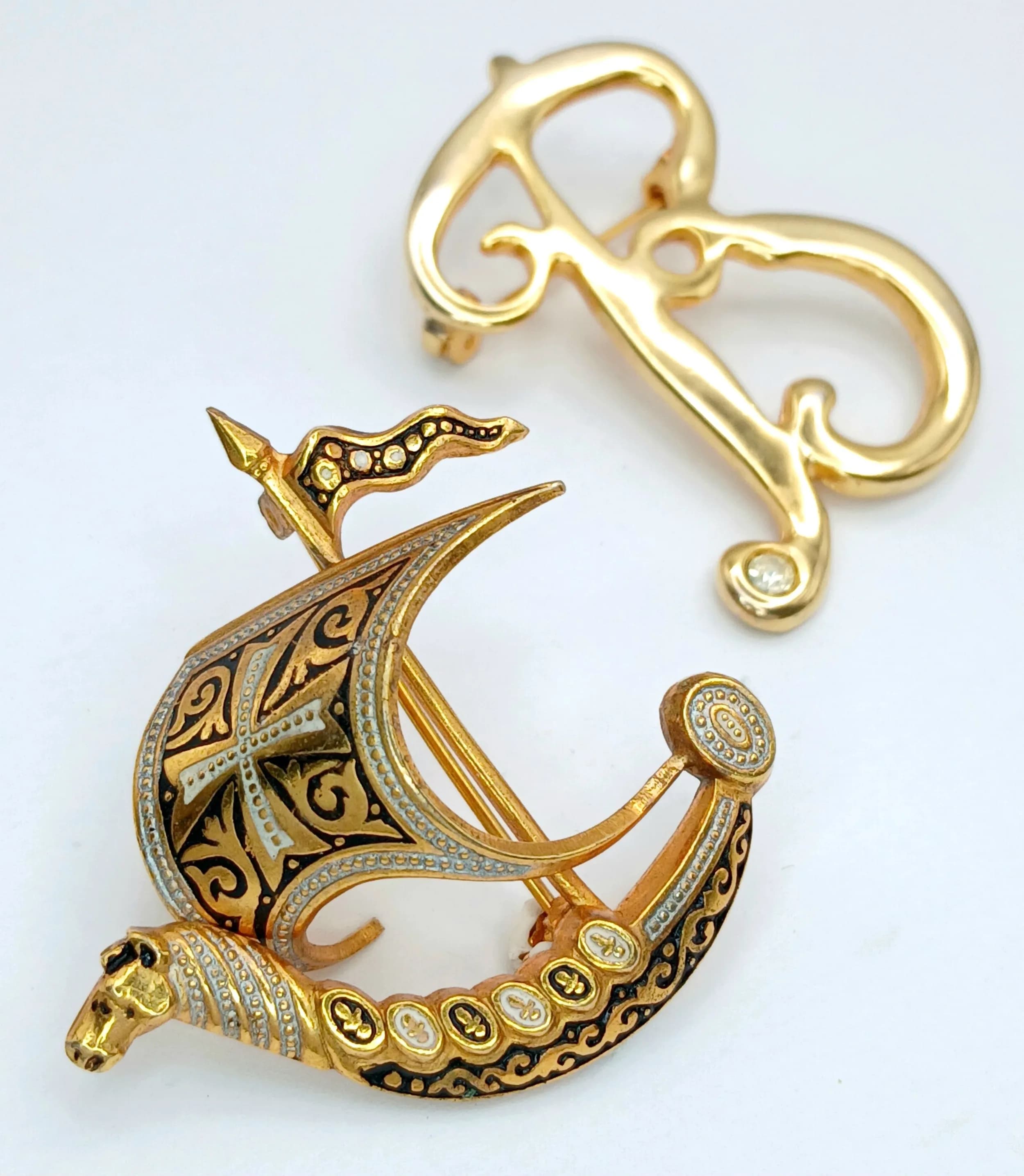 2x Lovely Gold Tone Brooches. Comprising an ornate initial “B” brooch and an enamelled Viking longship brooch. The initial brooch measures 4cm x 2.5cm. The ship brooch measures 4cm x 4cm.