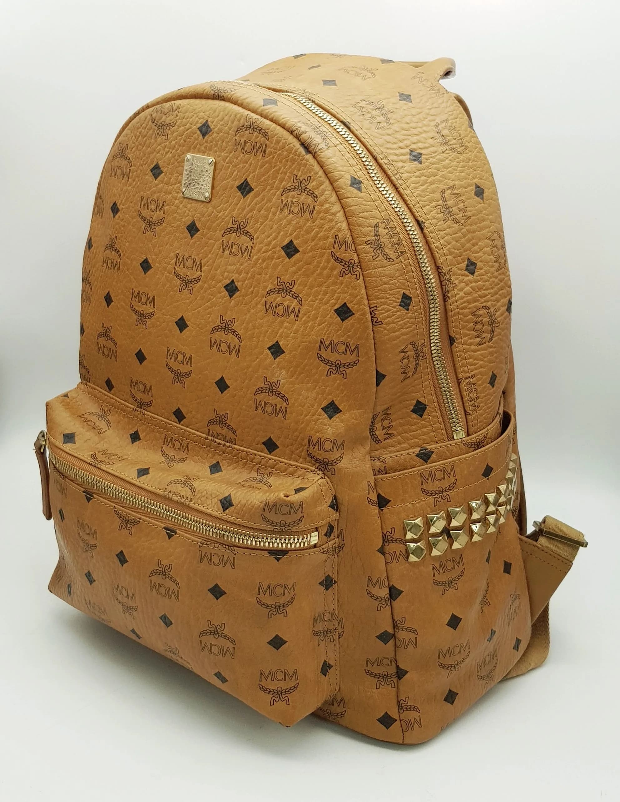 An MCM Visetos Large Stark Backpack. Cognac MCM monogram coated canvas exterior with gold-toned hardware, leather top handle, adjustable backpack straps, front zipped pocket and side pockets with stud detail. The zip-around closure opens to a black leather interior with one main compartment, an open inner side pocket and a padded compartment. Comes with dust cover bag. Approx 33cm length x 40cm height x 14.5cm depth. Some light wear to the corners, light scratches to the metal hardware and some stains to the interior. Please see photos or request a condition report. Ref: 26620.