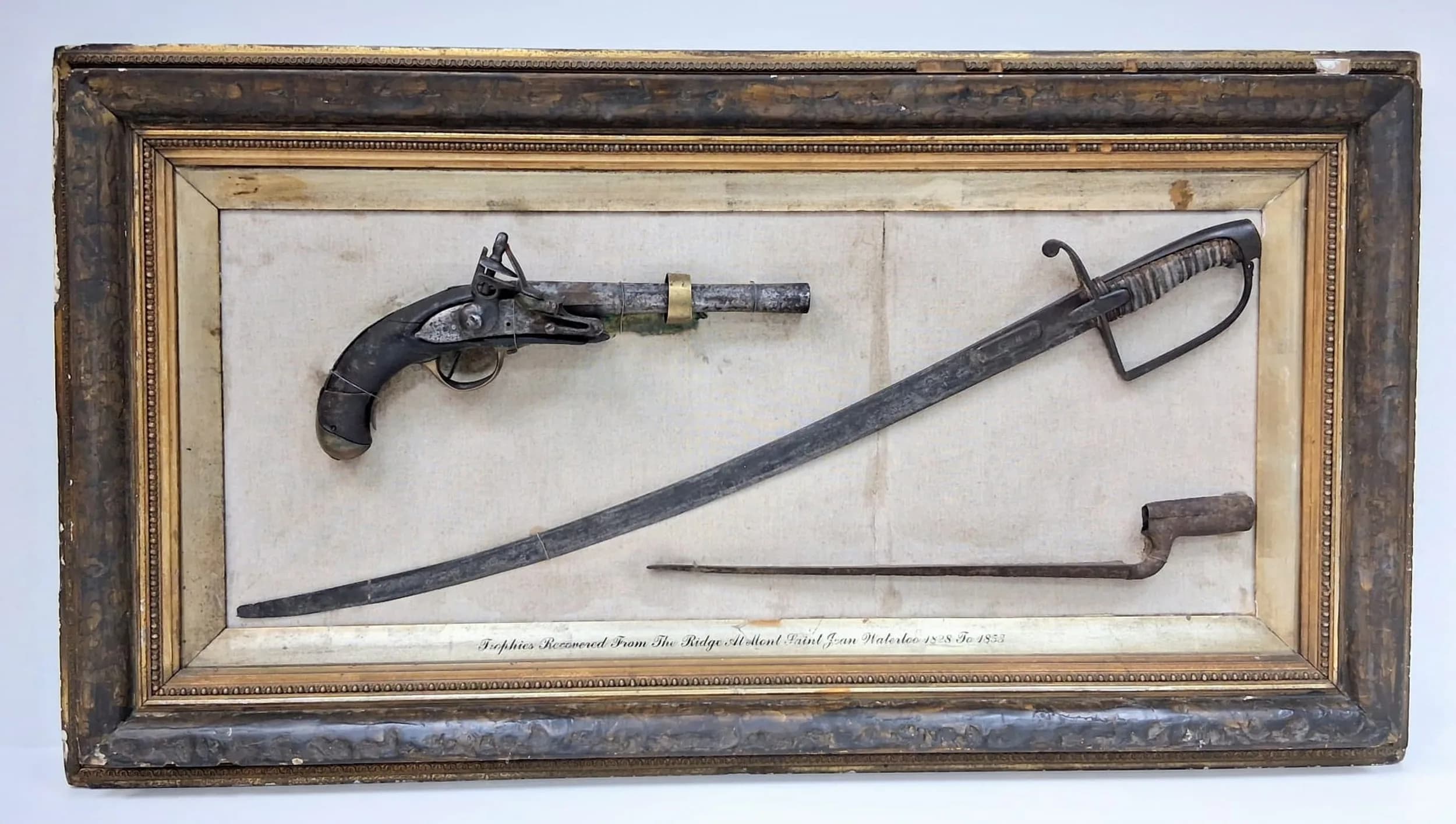 A Battle of Waterloo Relic Board - Containing Trophies Recovered From The Ridge At Mont Saint-Jean, Waterloo, Between The Years of 1828 -1853. Displayed in the Sergeant's Mess of the 73rd Regiment of Foot. The period frame contains three items - A French Cavalry Pistol, A British Officers Cavalry Sword and a British Brown Bess Socket Bayonet. Sergeant Mess 73rd Foot - Hand Written to Top Right Hand Corner Verso. 105x 56cm. UK Mainland Shipping Only