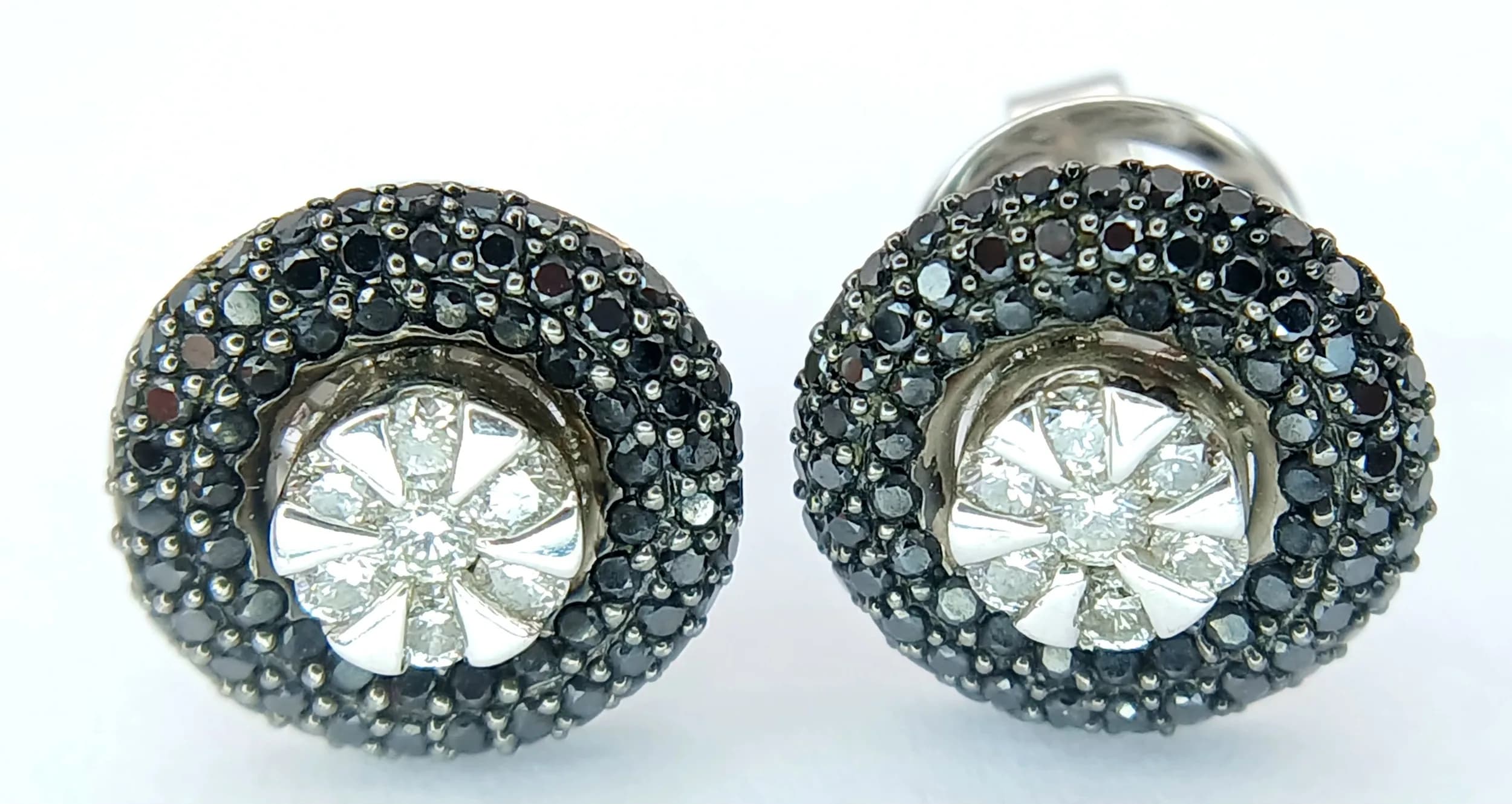 A Pair of 18K White Gold, Black and White Diamond Stud Earrings. Wheel form with 0.28ctw of white inner diamonds surrounded by a cluster of black diamonds. White diamonds having G colour and VS2 clarity. 5.2g total weight. MT27.