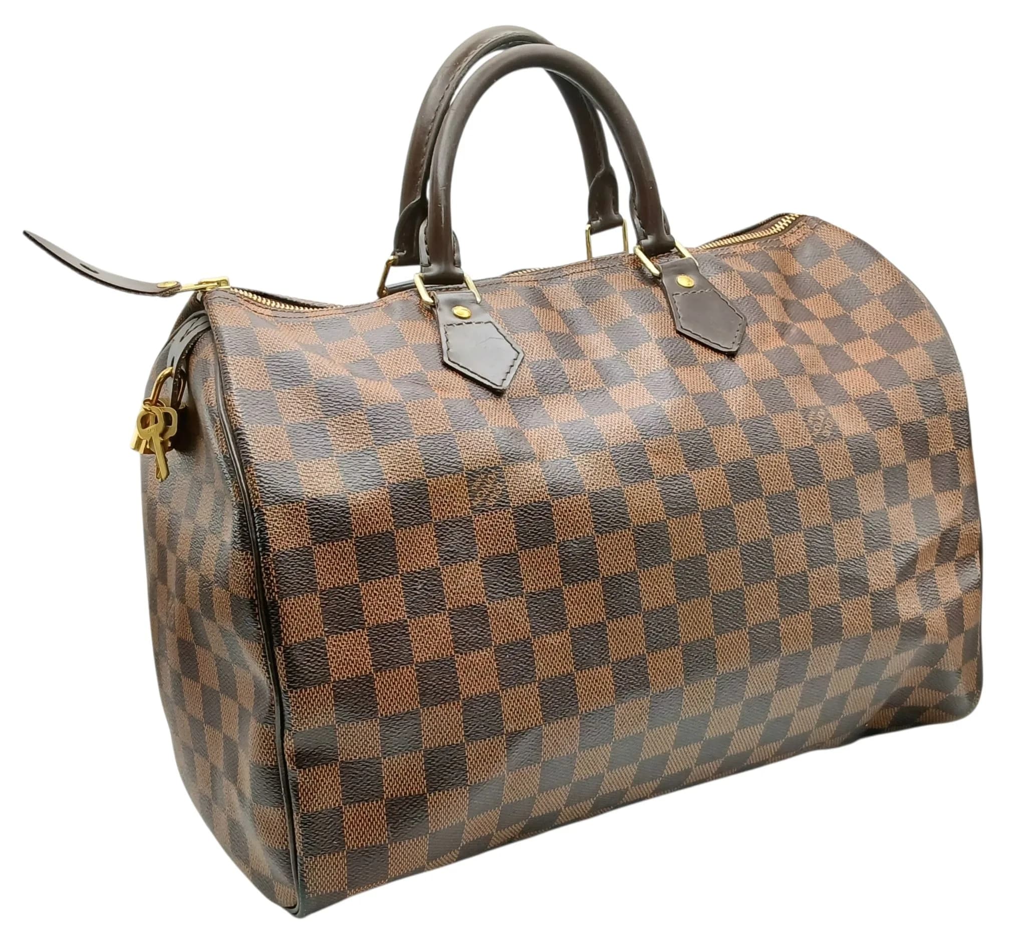 A Brown Louis Vuitton Damier Ebene Speedy 35 Bag. Brown Damier Ebene coated canvas exterior with gold-tone hardware, rolled top handles and top zip closure. Red textile interior with one main compartment and a slip pocket. Approx. 35cm length x 25cm height x 19cm depth. Comes with keys and padlock. Some marks, signs of wear and tear to the leather, scratches to the hardware. Please see photos or request a condition report. Ref: 29318.