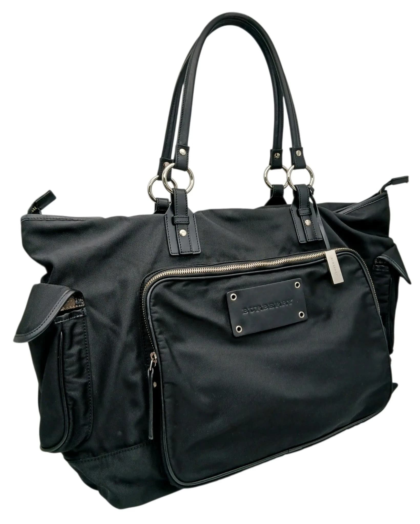 A Burberry Black Nylon Travel Holdall Bag. Black nylon exterior with silver-toned hardware, dual top handles and zipped top closure. Features one zipped pocket at the front and two flap pockets on both sides exterior. Signature Burberry Nova Check lining interior with one main compartment, an internal zipped pocket and opened pocket. Approx. 50cm length x 30cm height x 12cm depth. Some marks and signs of wear to the textile. Please see photos or request a condition report. Ref: 29537.