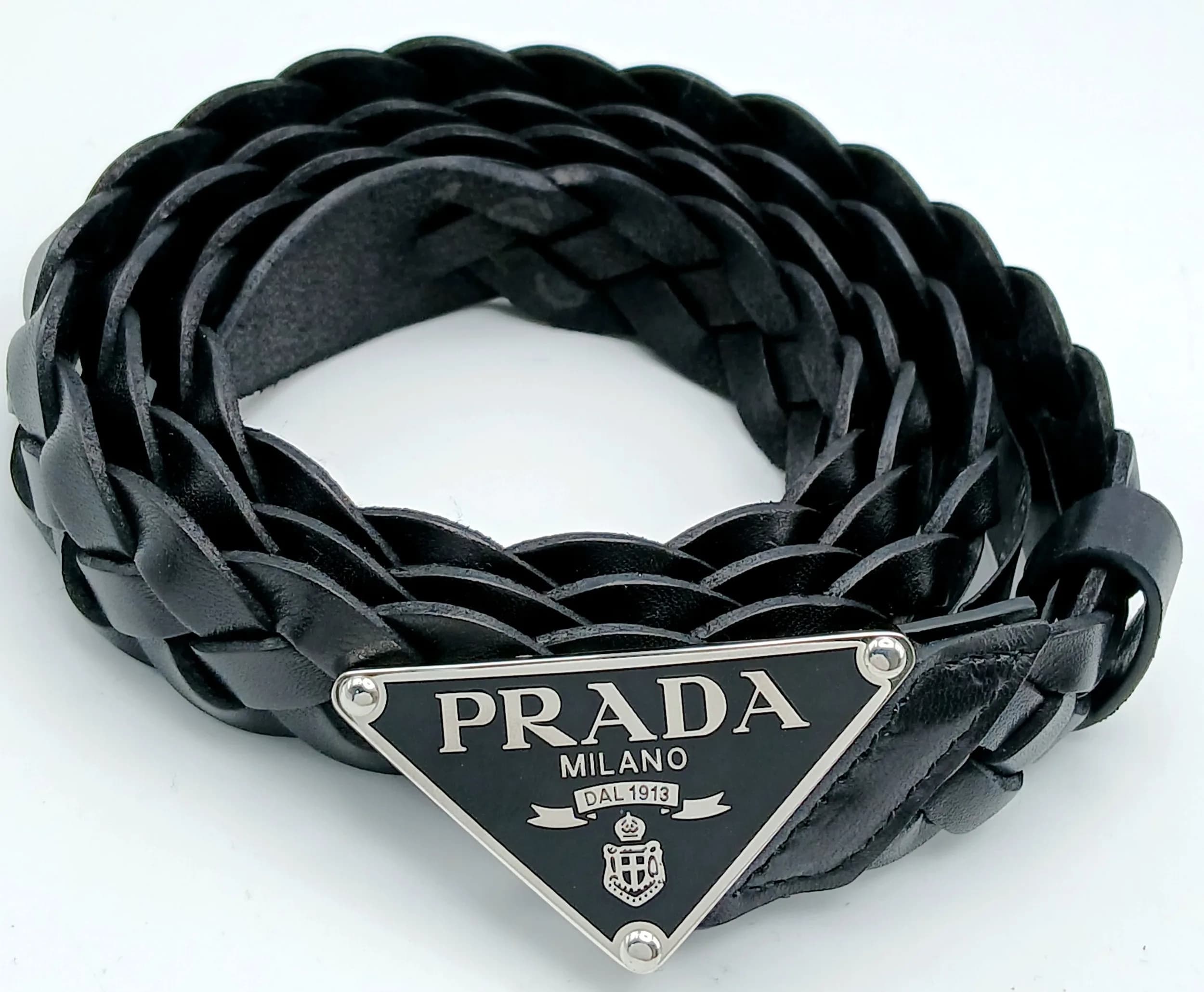 A Prada Black Braided Leather Belt. Black braided leather construction with a black and silver-tone Prada Milano triangle logo plaque to the front and adjustable buckle fastening. Approx. 113cm total length. Some marks and signs of use to the leather. Comes with a dust cover, RFID tag and care booklet, Please see photos or request a condition report. Ref: 29267.