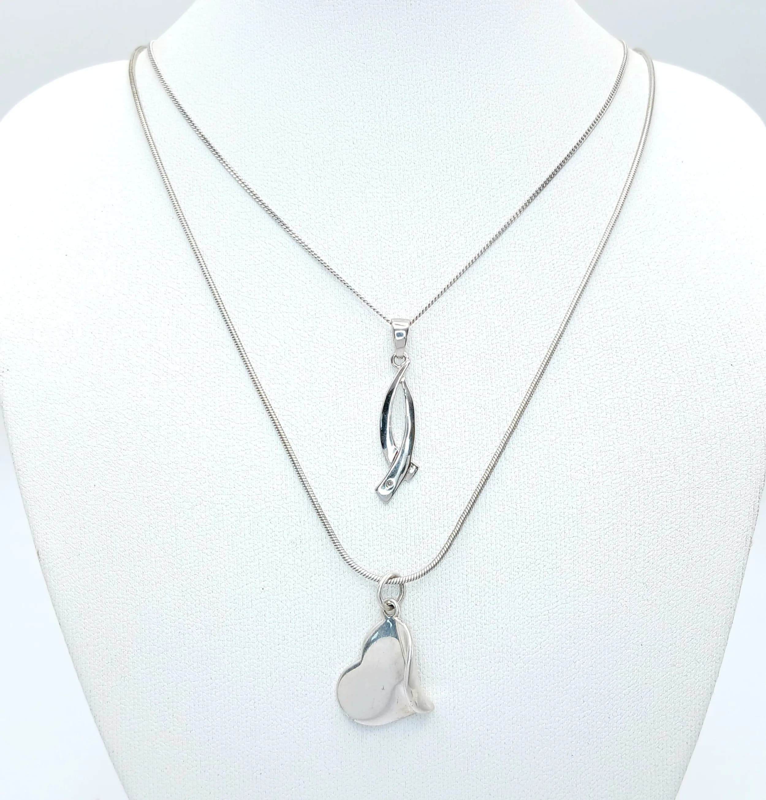 Two Sterling Silver Necklaces. Comprising: 1) A Very Unique Design Sterling Silver Heart Pendant Necklace-41cm Length Rope Chain. The Pendant measures a 2.5cm drop, 1.5cm width. Gross Weight 5.68 grams and 2) A White Sapphire Set Ribbon Design Pendant Necklace-46cm Length Chain. The Pendant measures a 3cm drop and is set with a round cut White Sapphire. Gross weight