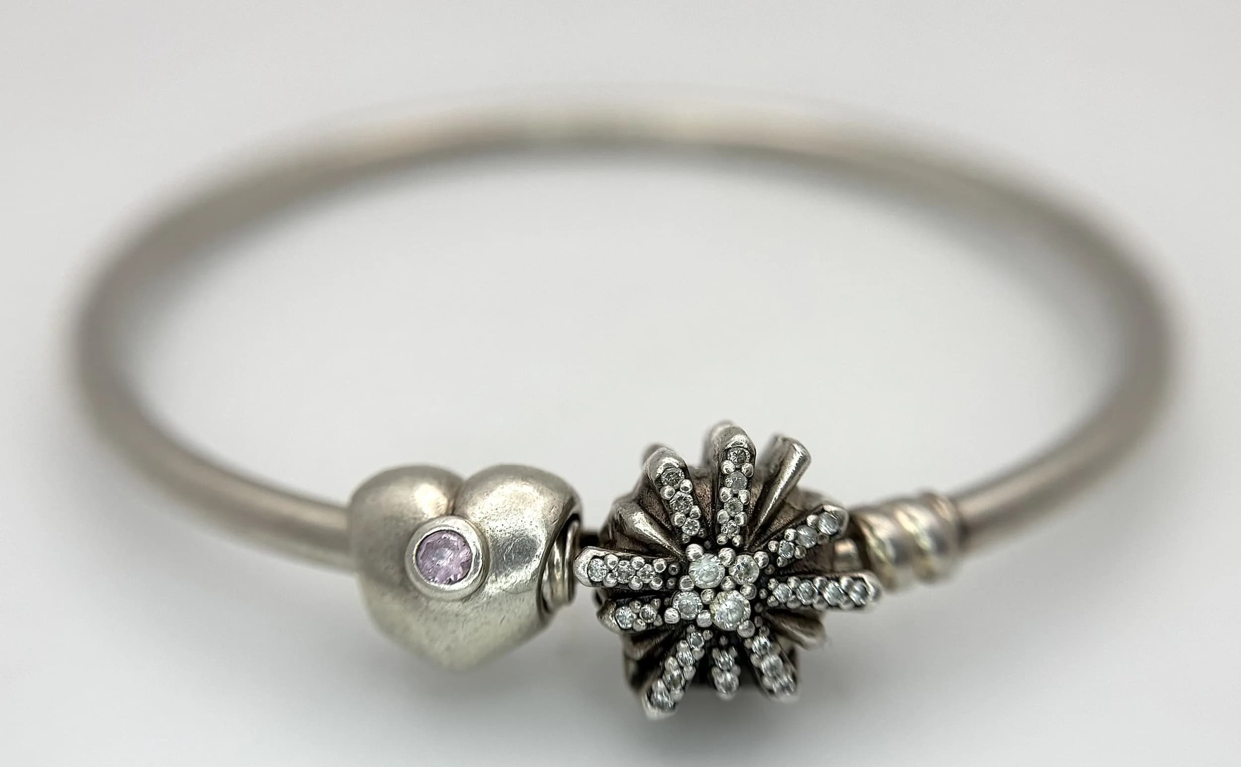 A Pretty Pandora 925 Silver Bangle Bracelet with Charms. Fitted with a polished silver bangle and two Pandora charms comprising a pink heart charm and a sparkling snowflake charm. The bangle measures approx 6cm internal diameter. Total weight 12.65 grams. Comes with a Pandora box.