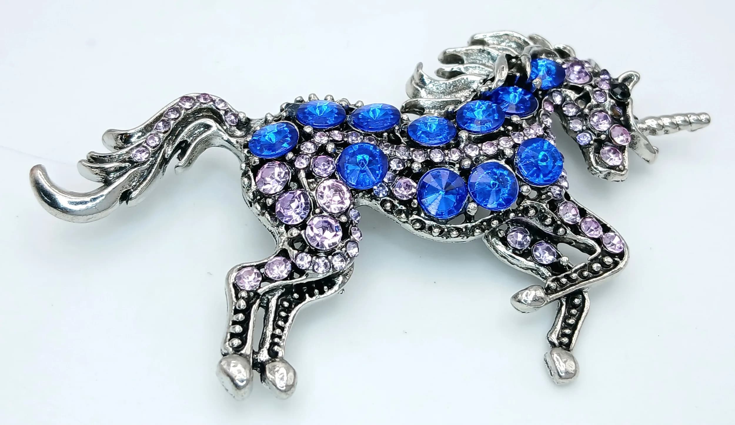 A Colourful Unicorn Stone Set Brooch. Set with round cut blue, pink and white stones in a silver tone mount, depicting a prancing unicorn. Measures approx 8.5cm x 5cm.