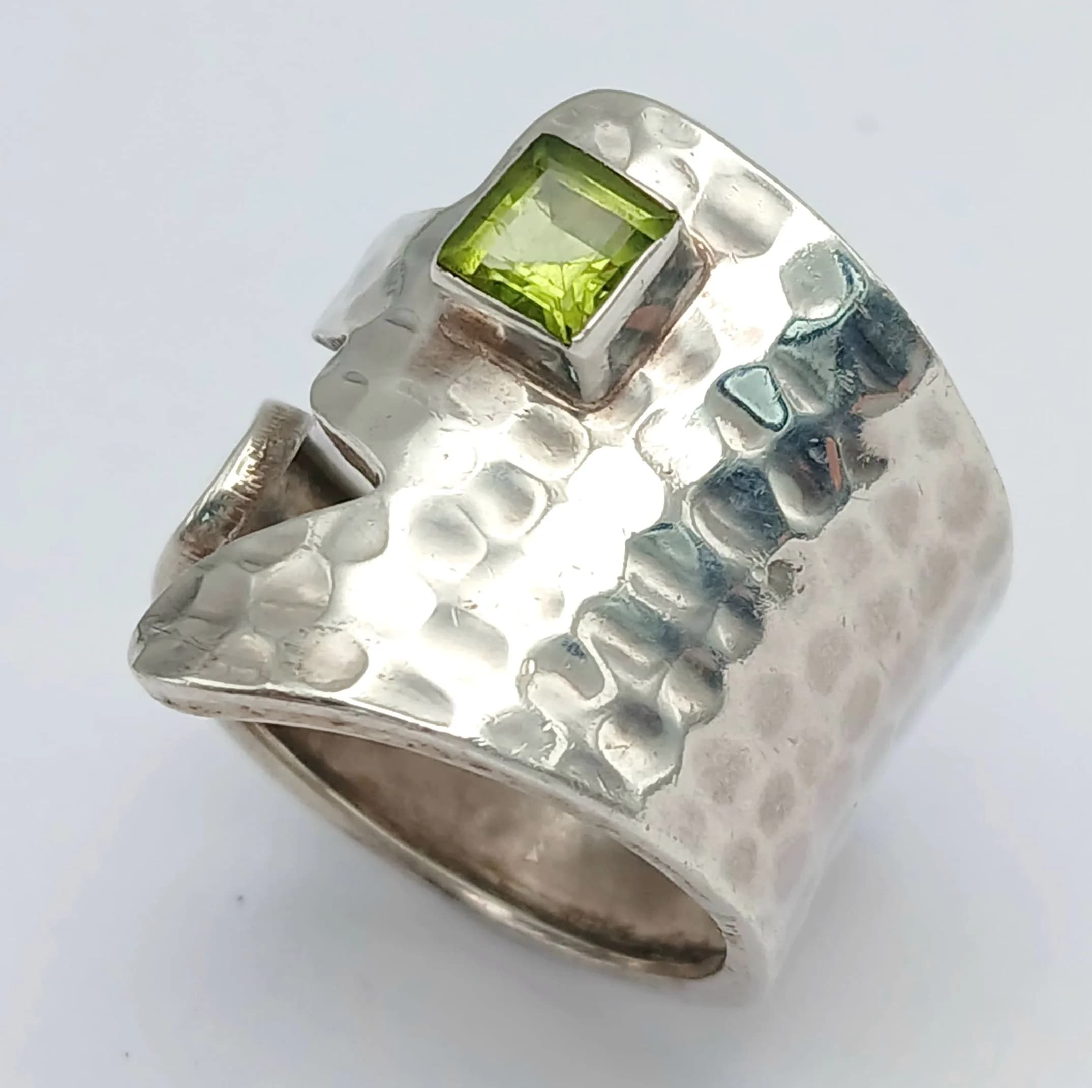 An Unique Unisex Sterling Silver Modernist Design Peridot Set Ring Size R. The Ring is set with a 5mm Square Cut Peridot, approximately 0.65 carat with the ring having a hammered finish. The Ring measures 2.3cm Wide at the Crown and has a gross weight of 7.91 grams