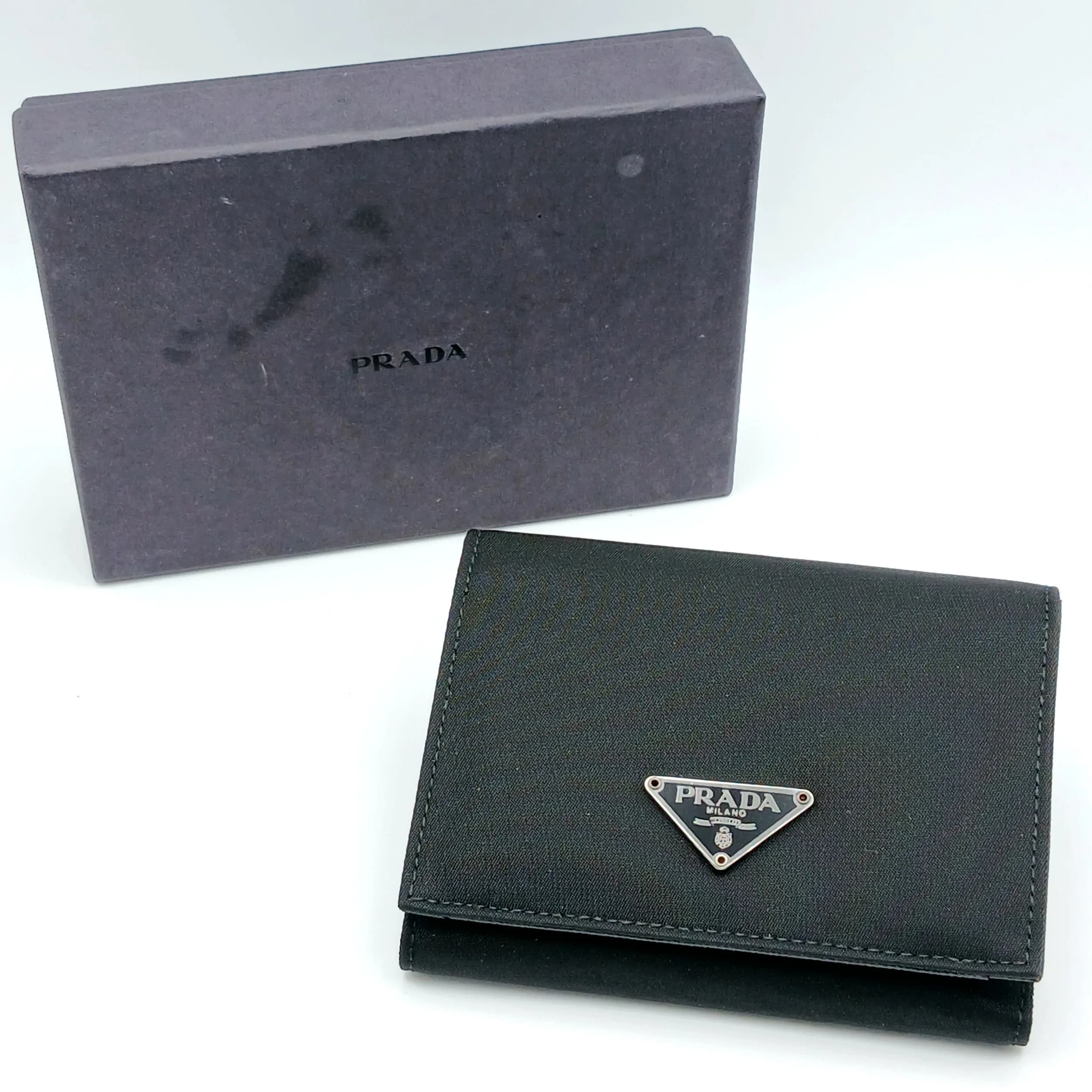 A Black Prada Leather Tri-Fold Wallet. Black leather exterior with silver-toned enamel Prada triangle logo to the front and press-stud fastening. Black leather interior with multiple card slots, note compartment and press-stud coin pocket. Approx. 11cm length x 10cm height (when closed). Comes with the Prada presentation box, tag and card of authenticity. Few marks and scratches but in very good condition. Please see photos or request a condition report. Ref: 29266.