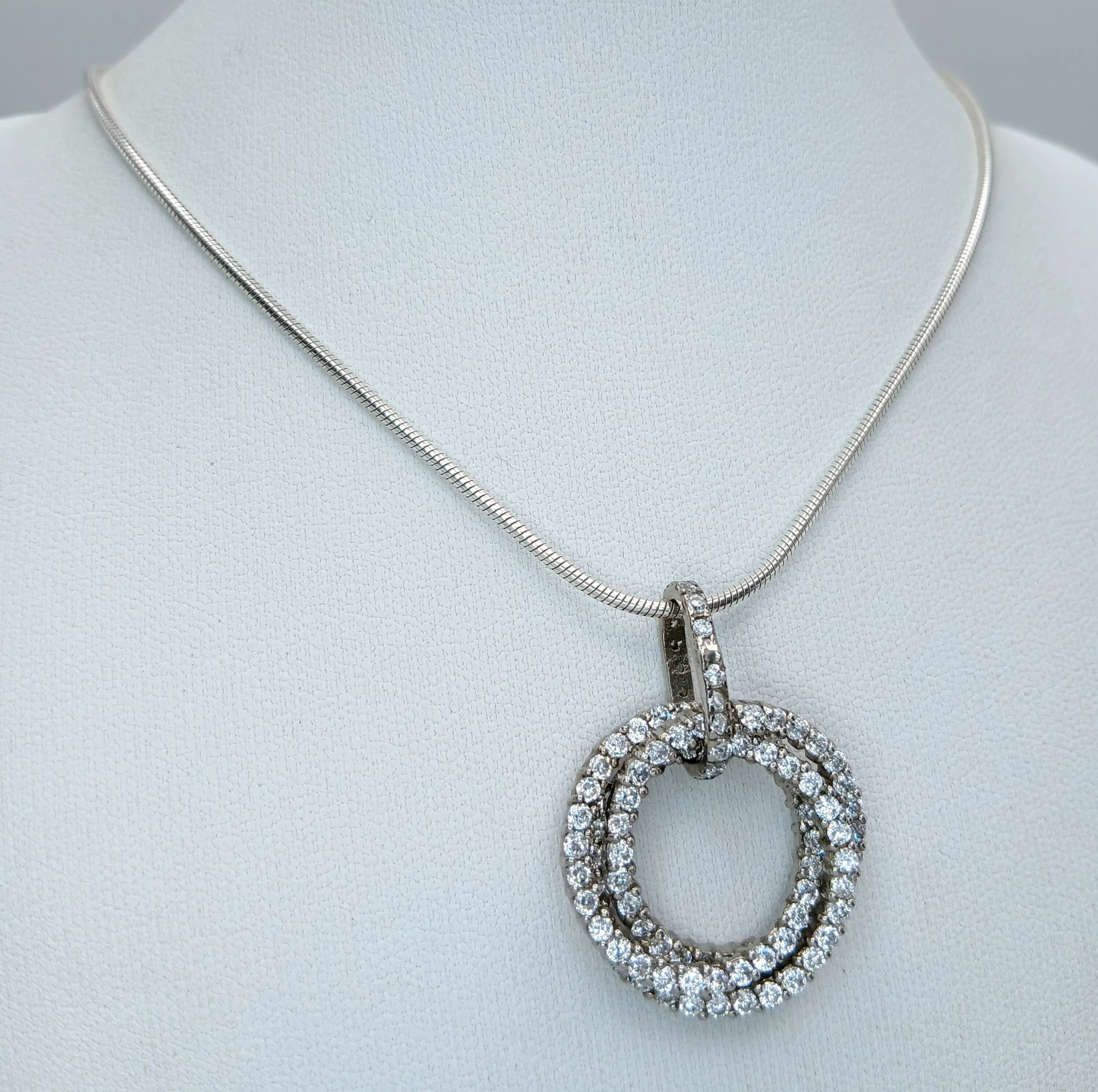 An Eye-catching 925 Sterling Silver CZ Set Circle Pendant Necklace - 36cm Length. Features an interlocking double circle pendant pave set with white CZ stones on a fine snake chain. The pendant measures 2.5cm diameter. Gross weight 12.08 grams.