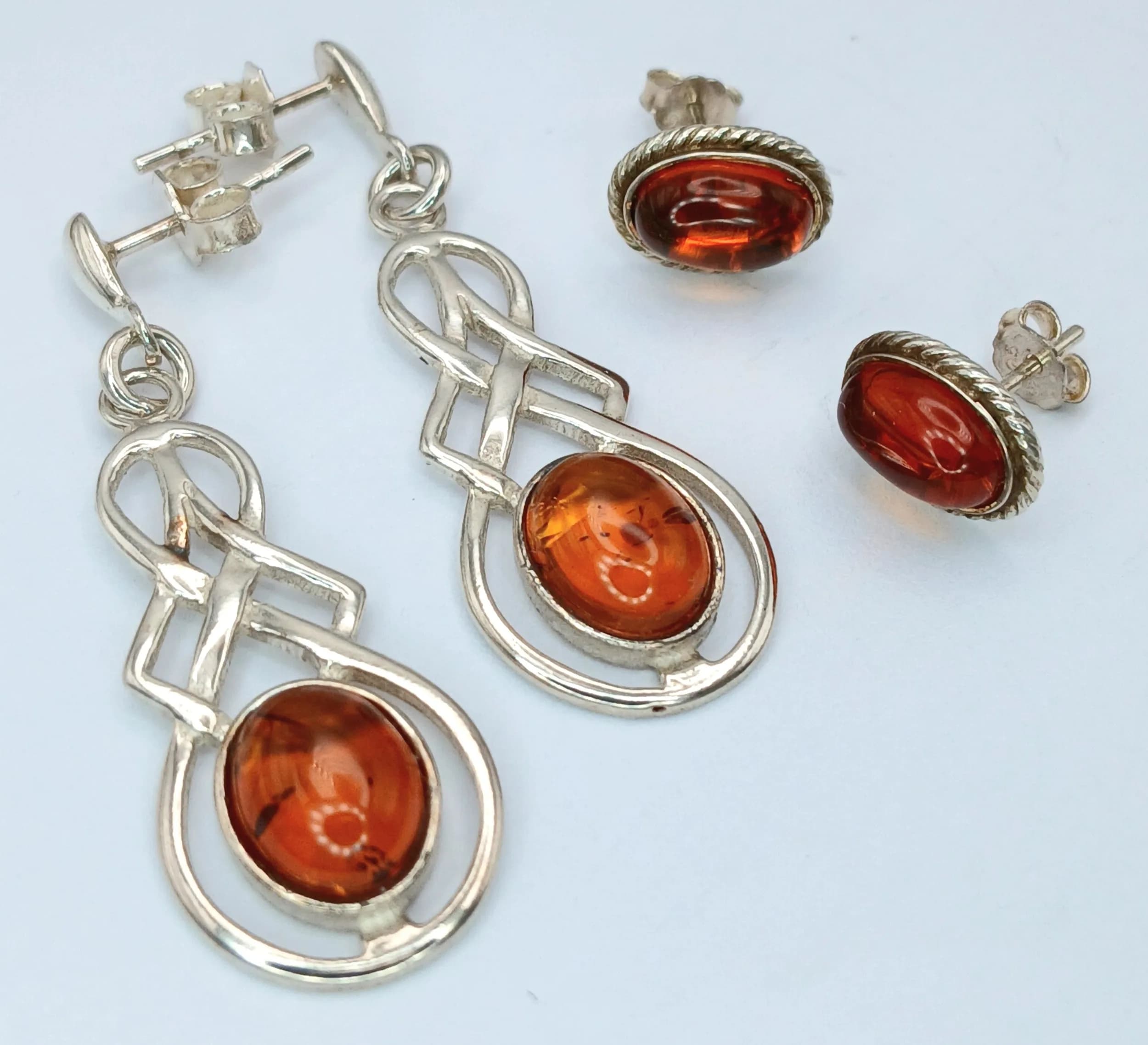 Two Pairs of Silver and Amber Earrings.. Comprising: 1) A Pair of Honey Coloured Oval Amber Cabochon Stud Earrings 1cm Length and 2) A Pair of Celtic Knot Design Cognac Coloured Oval Amber Dangle Earrings. 3.6cm Length.