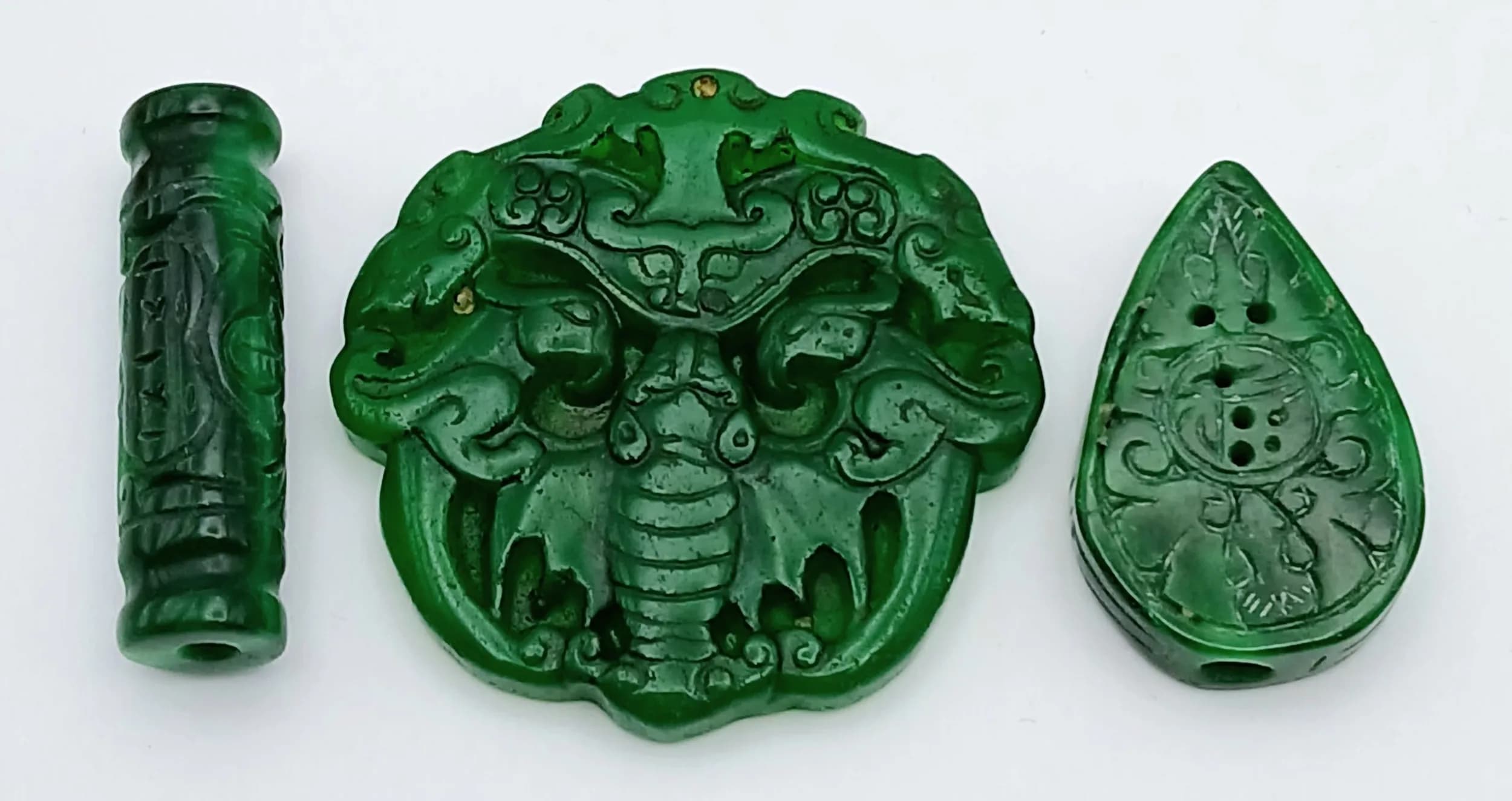 A Parcel of Three Intricately Carved Chinese Green Jade Pendants. The Largest Dragon Detail pendant measures 5.3 x 5.3cm, the Cylindrical Pendant measures 4.3 x 1.2cm and the Pear Cut Amulet Pendant measures 4 x 2.5cm.