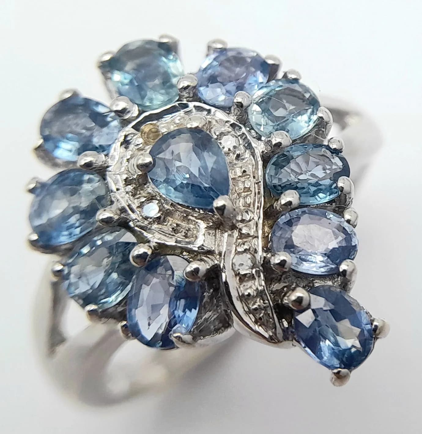 A Beautiful 9k White Gold Blue Sapphire and Diamond Set Ring. Size N. The Ring is set with a centre 4 x 3mm Pear Cut Tanzanian Blue Sapphire (0.142 carat), ten 4 x 3mm Oval Cut Cambodian Blue Sapphires (1.88 ctw) and four1mm round African Diamonds (0.02 ctw). The Ring has a gross weight of 5 grams. Appears Practically Unworn if worn at all.