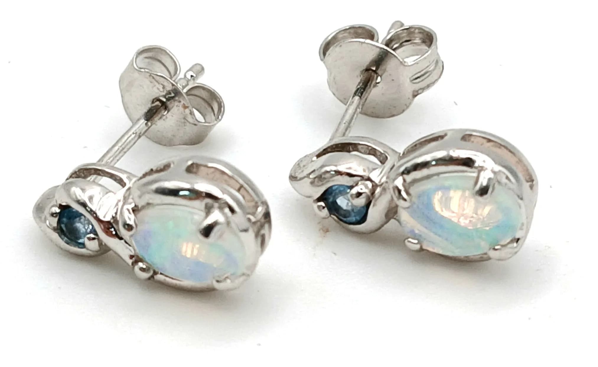 A Beautiful Pair of 9k White Gold Opal and Blue Topaz Stud Earrings 1.2cm Length. Each Earring is set with a 6 x 4mm Oval Opal, circa 0.30 carat with Round Cut Blue Topaz Set above. The Earrings have a combined gross weightof 1.8 grams.