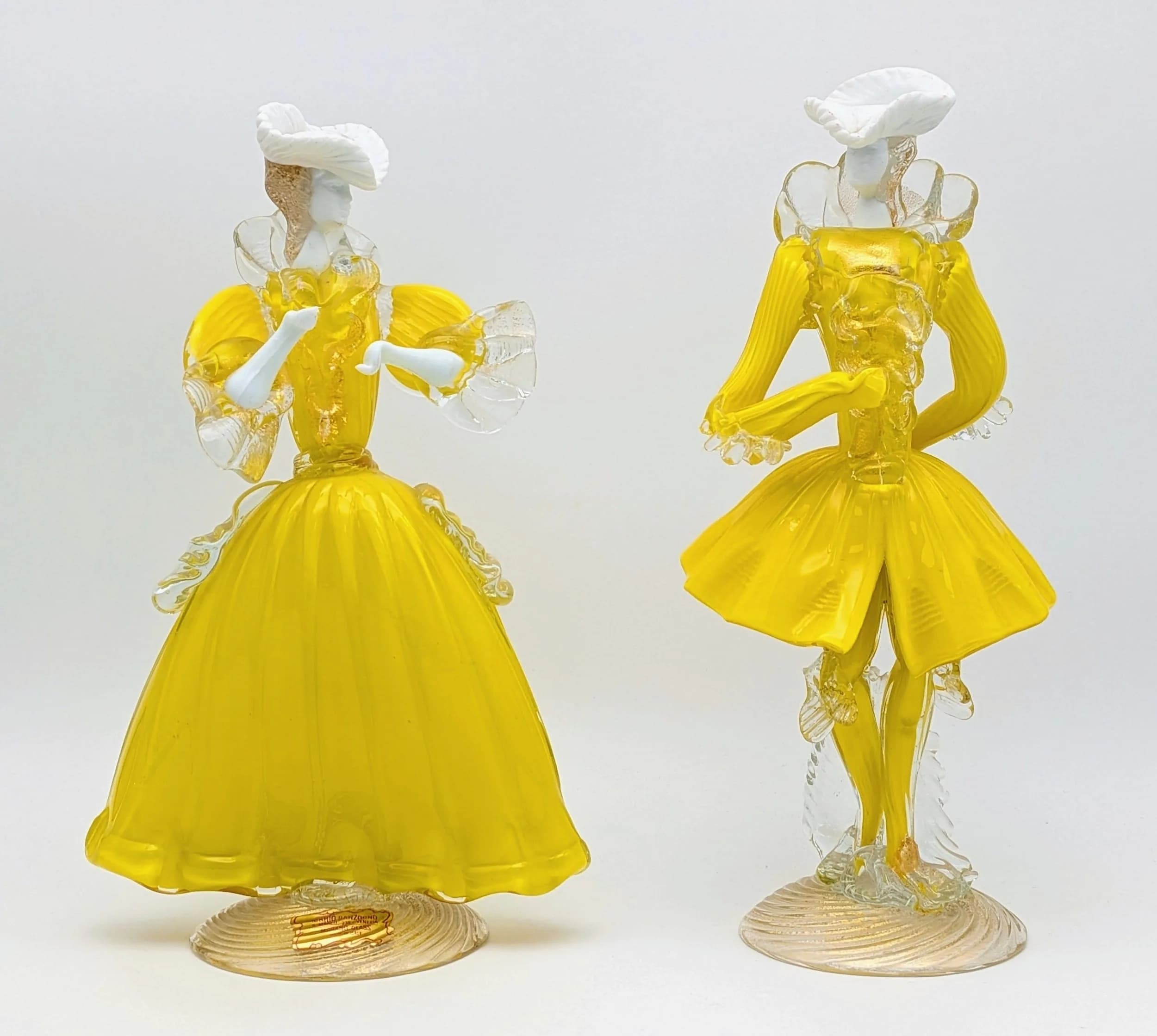 A stunning pair of vintage Murano glass Venetian figures. Depicting a couple of traditional Venetian 'Dandy' carnival dancers. Hand blown in striking yellow with white details and gold dust (Aventurine) inclusions. Very good condition. No chips or cracks. One still has the label for the prestigious Mario Sanzogno Gallery. 24cm tall.