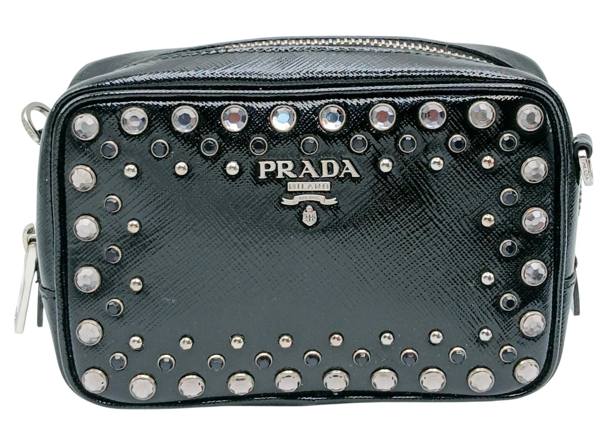 A Prada Studded Saffiano Leather Camera Crossbody Bag. Black patent-finish Saffiano leather exterior embellished with silver-tone studs and faceted crystal detailing, with Prada Milano logo plaque to the front. Zip-around closure with Prada-branded silver-tone hardware and a detachable black leather shoulder strap. Black Prada jacquard interior with an open inner pocket and card slots. Approx. 15cm length x 10cm height x 5.5cm depth. Comes with Prada dust bag and certificate of authenticity. In good condition overall. Please see photos or request a condition report. Ref: 28560.