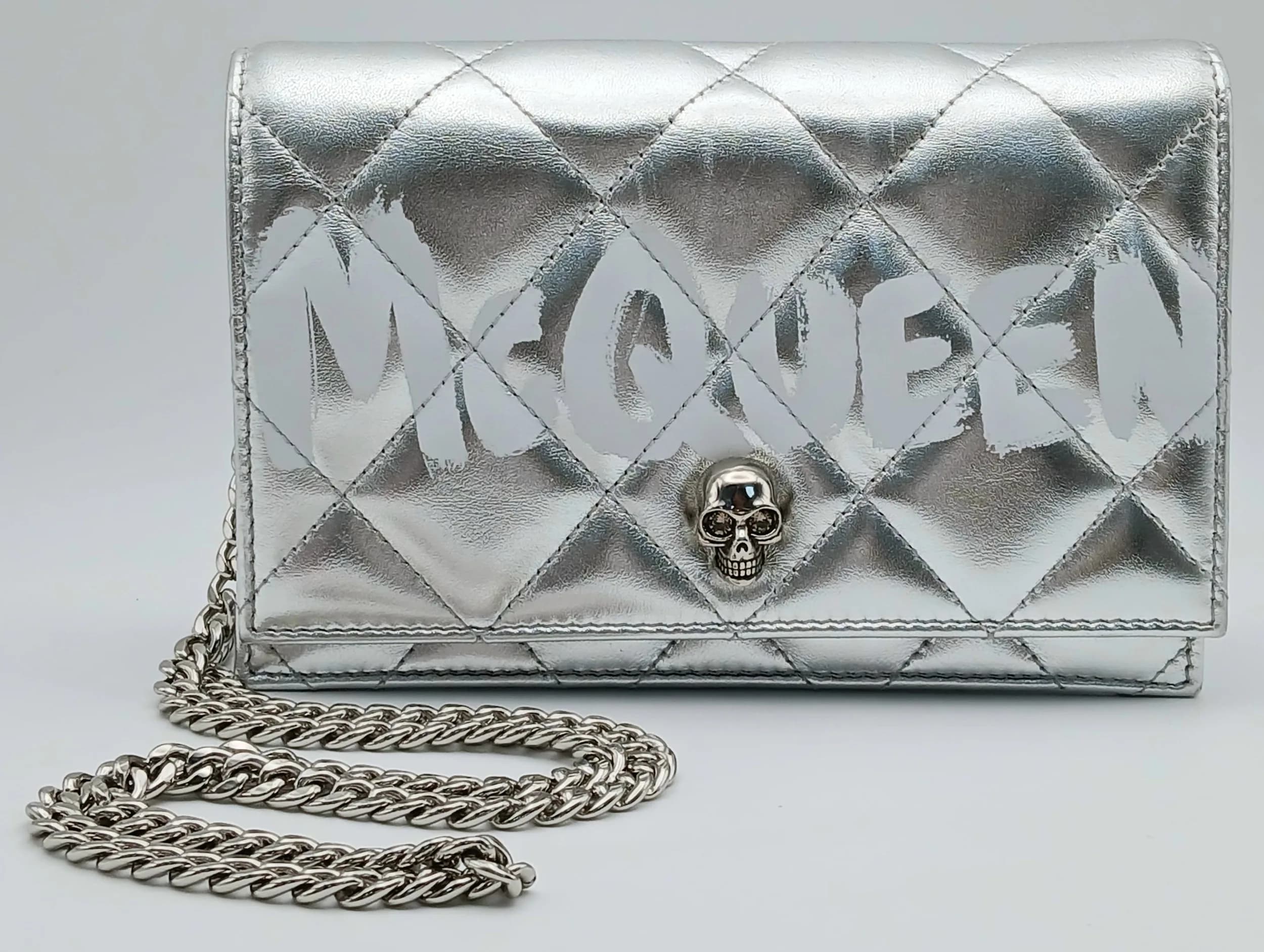 An Alexander McQueen Graffiti Small Skull Crossbody Bag. Silver metallic quilted leather exterior with white McQueen graffiti print, silver-tone hardware, signature skull embellishment and a silver-tone chain shoulder strap. Fold-over flap with magnetic press-stud closure. Black suede interior with one main compartment and an inner card pocket. Approx. 19.5cm length x 13.5cm height x 5cm depth. Comes with a dust cover bag. In good condition overall. Please see photos or request a condition report. Ref: 26901.