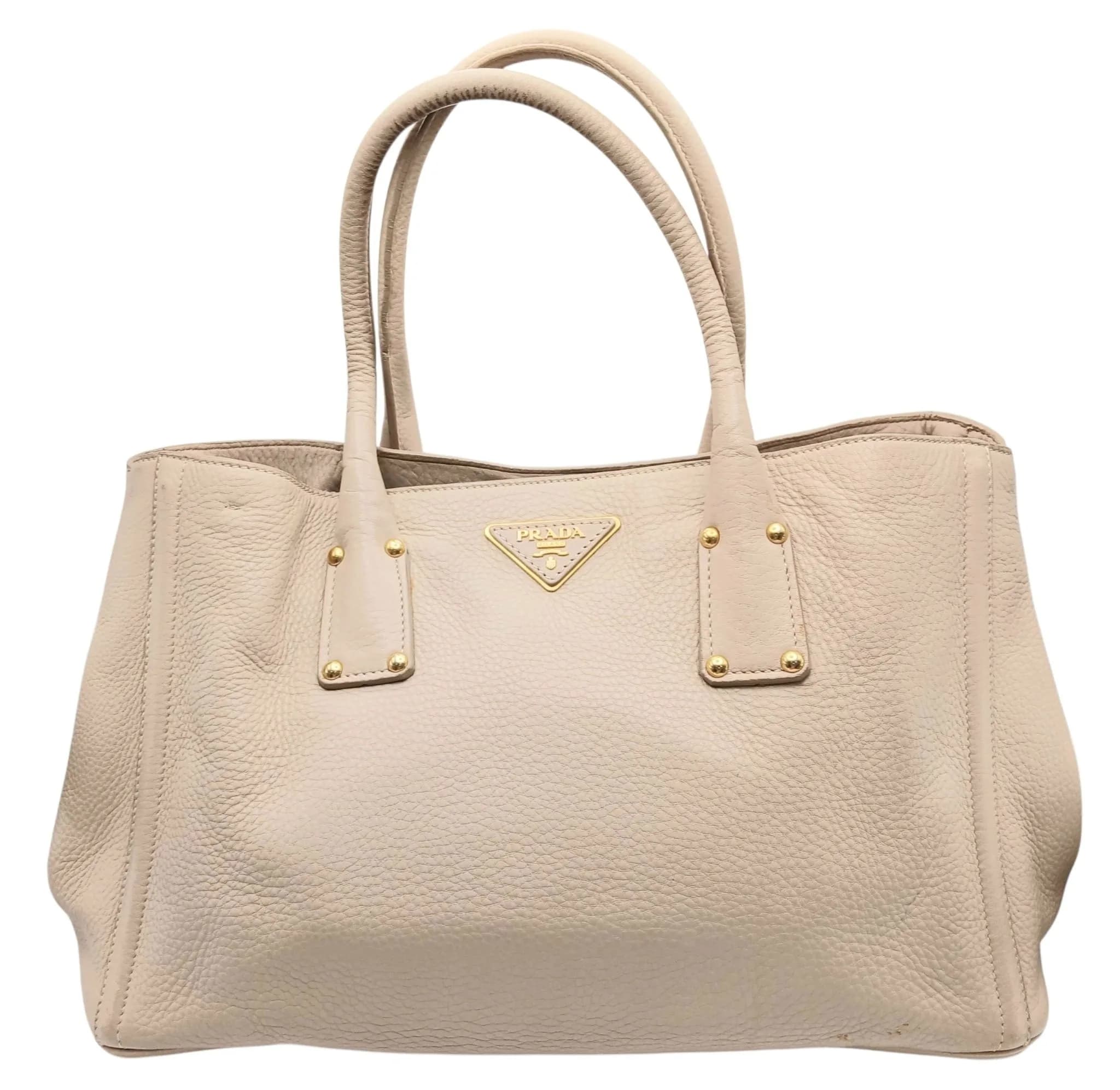 A Prada Vitello Daino Leather Tote Bag. Beige pebbled leather exterior with gold-tone hardware, Prada triangle logo plaque, double leather top handles, press-stud closures and four protective base studs. Beige textile interior with one main compartment, two zipped inner side pockets and two open inner side pockets. Comes with a detachable leather shoulder strap. Approx. 43cm length x 25cm height. Signs of wear to the corners and stains to the exterior. Pen marks and stains to the interior, with some fraying and wear to the interior fabric - Please see photos or request a condition report. Ref: 29776.