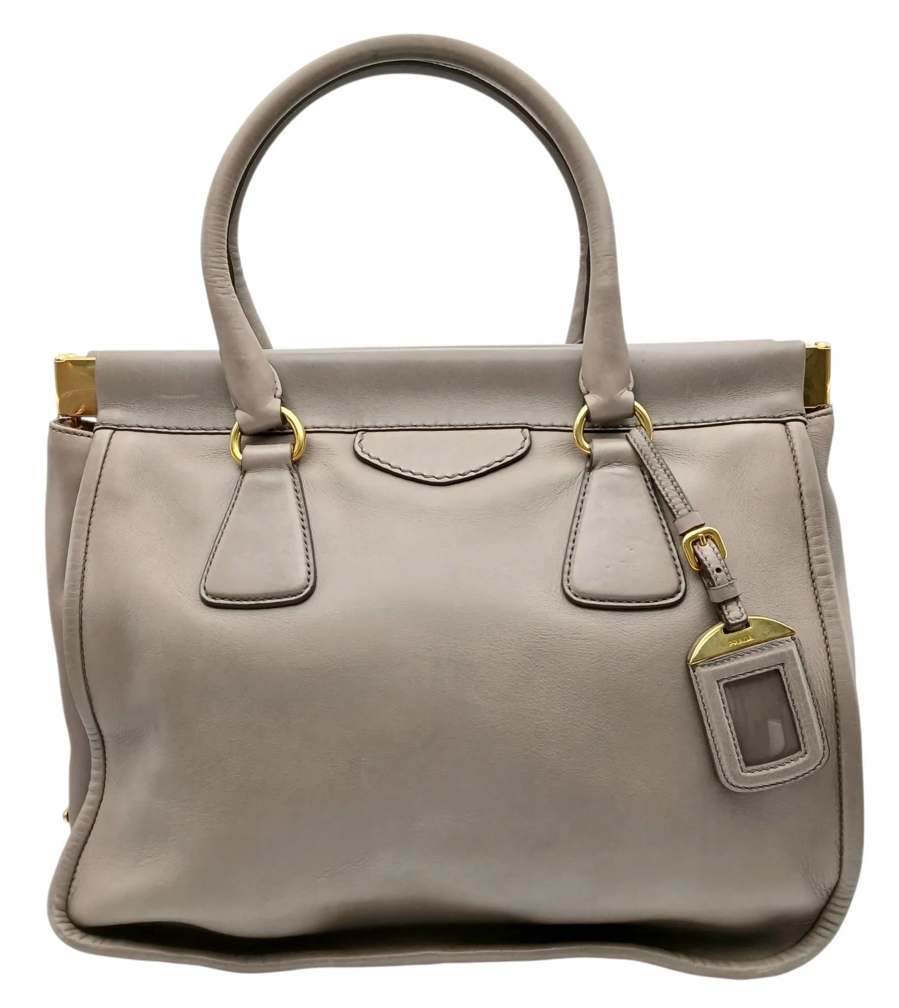 A Prada Taupe Leather Tote Bag. Taupe leather exterior with gold-tone hardware, double rolled leather top handles, structured frame top and Prada branded leather luggage tag. Opens to a beige leather interior with two main compartments, one zipped inner pocket, one press-stud pocket and three open inner side pockets. Protective gold-tone base studs. Approx. 35cm length x 27cm height. Some signs of wear to the corners and light creasing to the leather exterior - Please see photos or request a condition report. Ref: 29780.