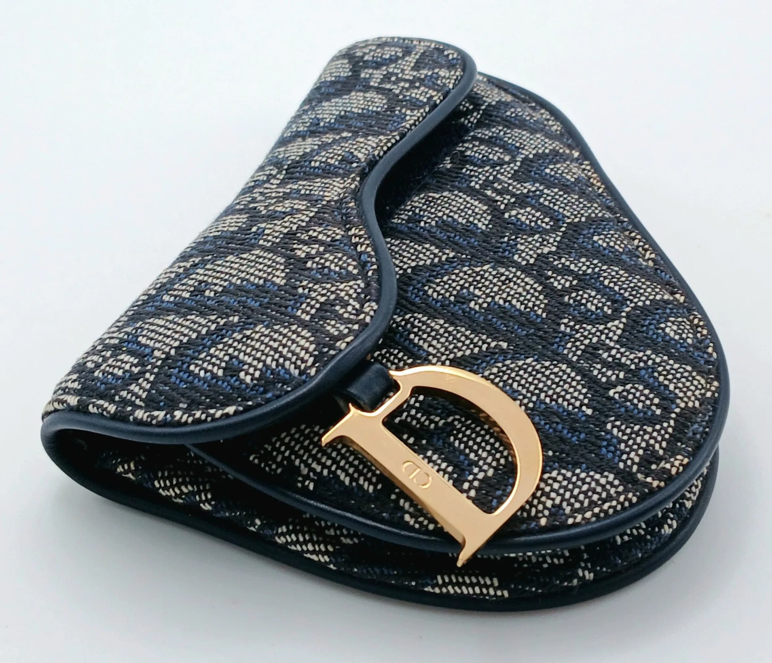 A Christian Dior Navy Blue Oblique Saddle Coin Purse. Navy blue and cream Dior Oblique jacquard exterior with navy leather trim and gold-tone ‘D’ chain hardware. Velcro flap closure opens to a navy textile interior with a zipped coin compartment and an open compartment. Approx. 13cm length x 9.5cm height. Overall in good condition. Please see photos or request a condition report. Ref: 29685.