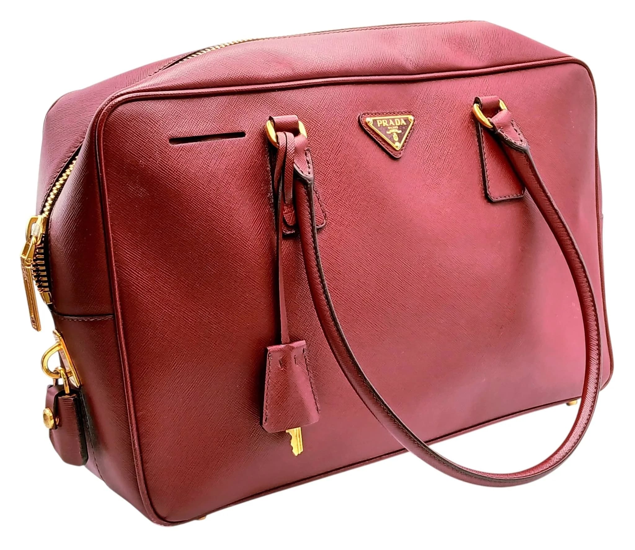A Prada Two Way Saffiano Leather Handbag. Burgundy Saffiano leather exterior with gold-tone hardware, double rolled leather top handles, top zip closure, Prada triangle logo plaque, protective base studs and clochette with keys. Opens to a dark brown Prada logo-jacquard textile interior with one main compartment and a zipped inner side pocket. Approx. 35cm length x 22.5cm height x 9.5cm depth. Signs of wear to the corners, light white marks to the exterior leather and signs of tarnishing to the zipper hardware - Please see photos or request a condition report. Ref: 29778.