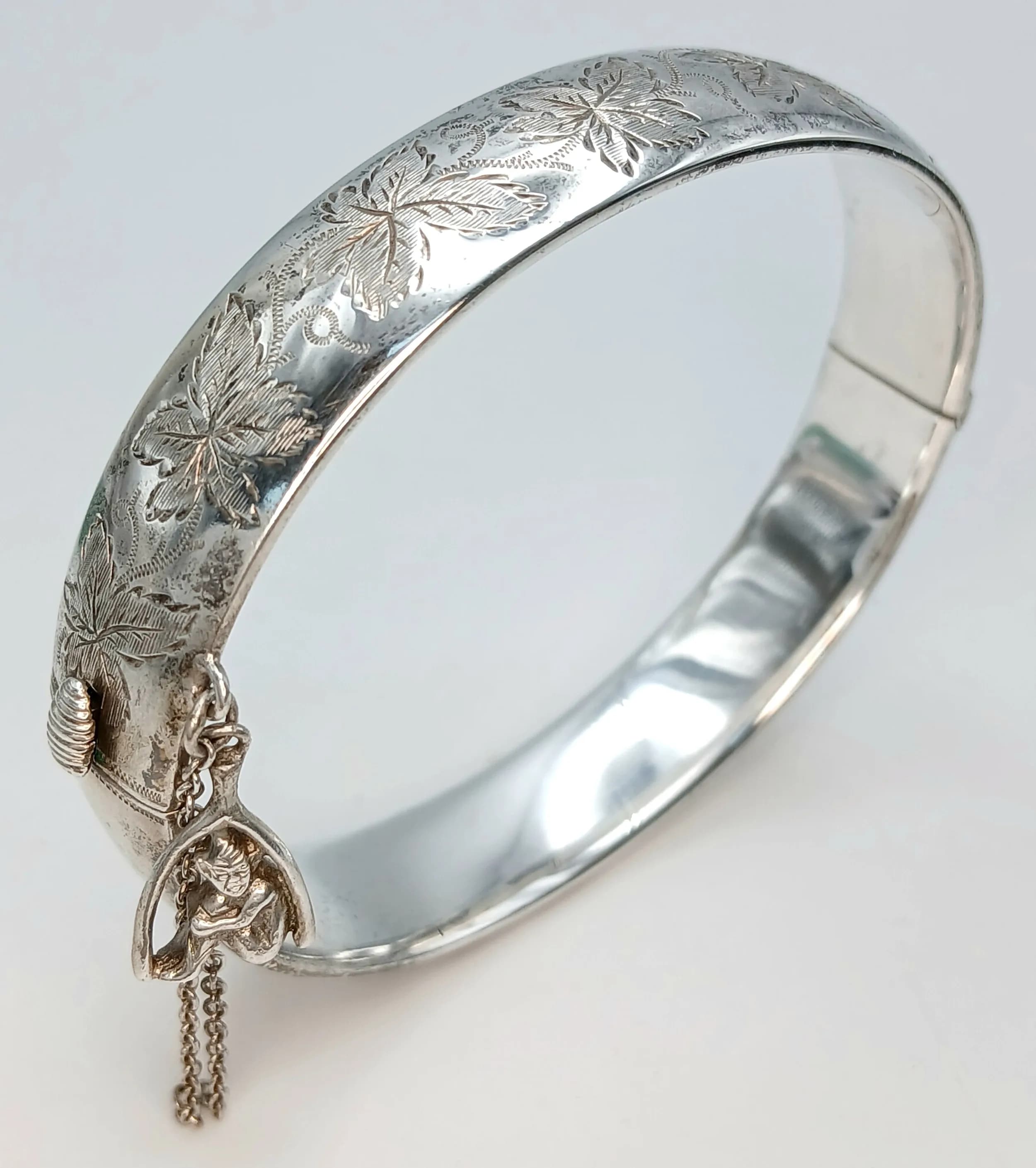 A SOLID SILVER HINGED BANGLE DECORATED WITH LEAF FIGURES TO ONE SIDE AND A PIXIE CHARM ATTACHED TO THE SAFETY CHAIN . 25.6gms