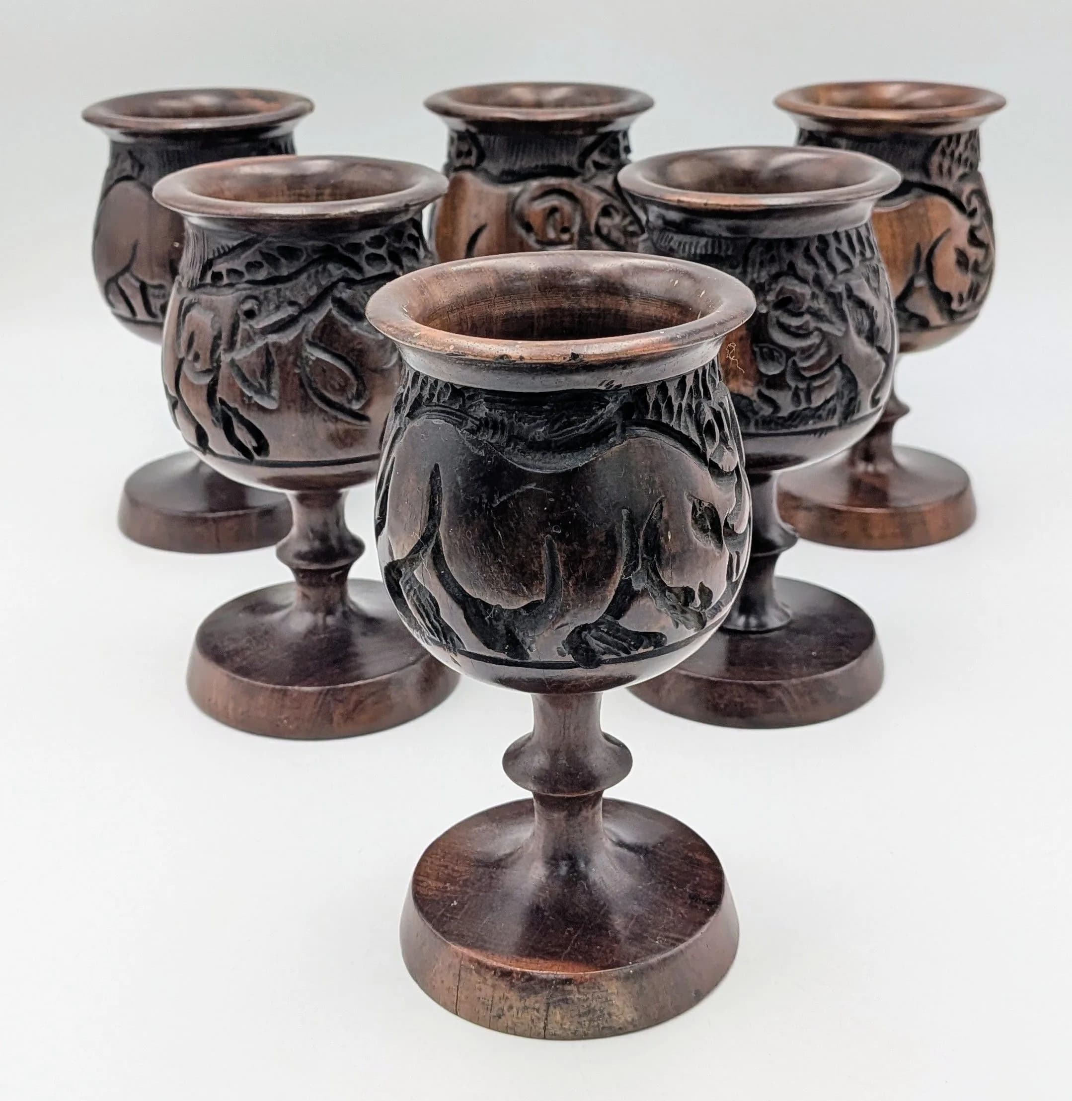 A Set of Six Carved Wooden Goblets. Each with carved rhinoceros and elephant decoration to the bowl, raised on circular pedestal bases. Approx 16cm height.