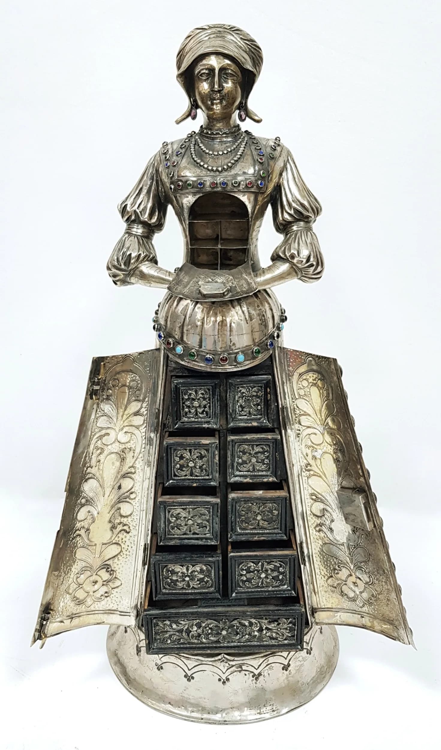 A Rare Antique German Late 19th Century, Silver and Stone Set Figural Jewellery Cabinet. Circa 1890, in the Renaissance 16th Century style. The large table cabinet modelled as a standing lady with headdress and gem set earrings. The dress is set with various semi-precious stones and hardstone cabochons, including turquoise, amethyst, lapis lazuli, garnets and agate. The front of the bodice is hinged and opens up to five compartments. The skirt of the dress has a pair of hinged doors that open to reveal a fitted interior of nine wooden drawers with silver acanthus leaf mounts with pseudo earlier marks. The interior of a drawer has a label inscribed ‘SAMMLUNG OTTO F. ERNST WIESBADEN No. 159’ with an old German catalogue entry and a hand written note regarding the pseudo marks. Approximately 6490g/208 oz/18 dwt weight. 72cm tall. Purchased at Sotherby's. Winning bidder must collect from Cadmore HQ or arrange a courier service.